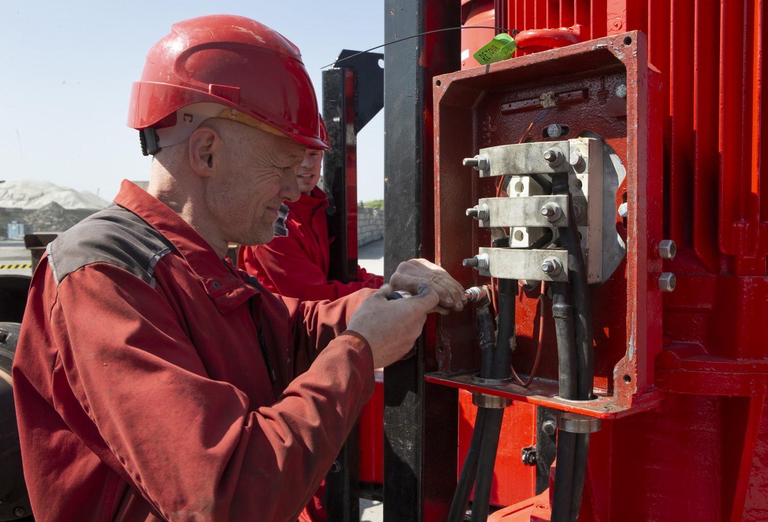Elektromonteur aan het werk aan een elektromotor Een monteur in rode overall en met een rode helm draait een moer vast in een elektrische installatie met kabels