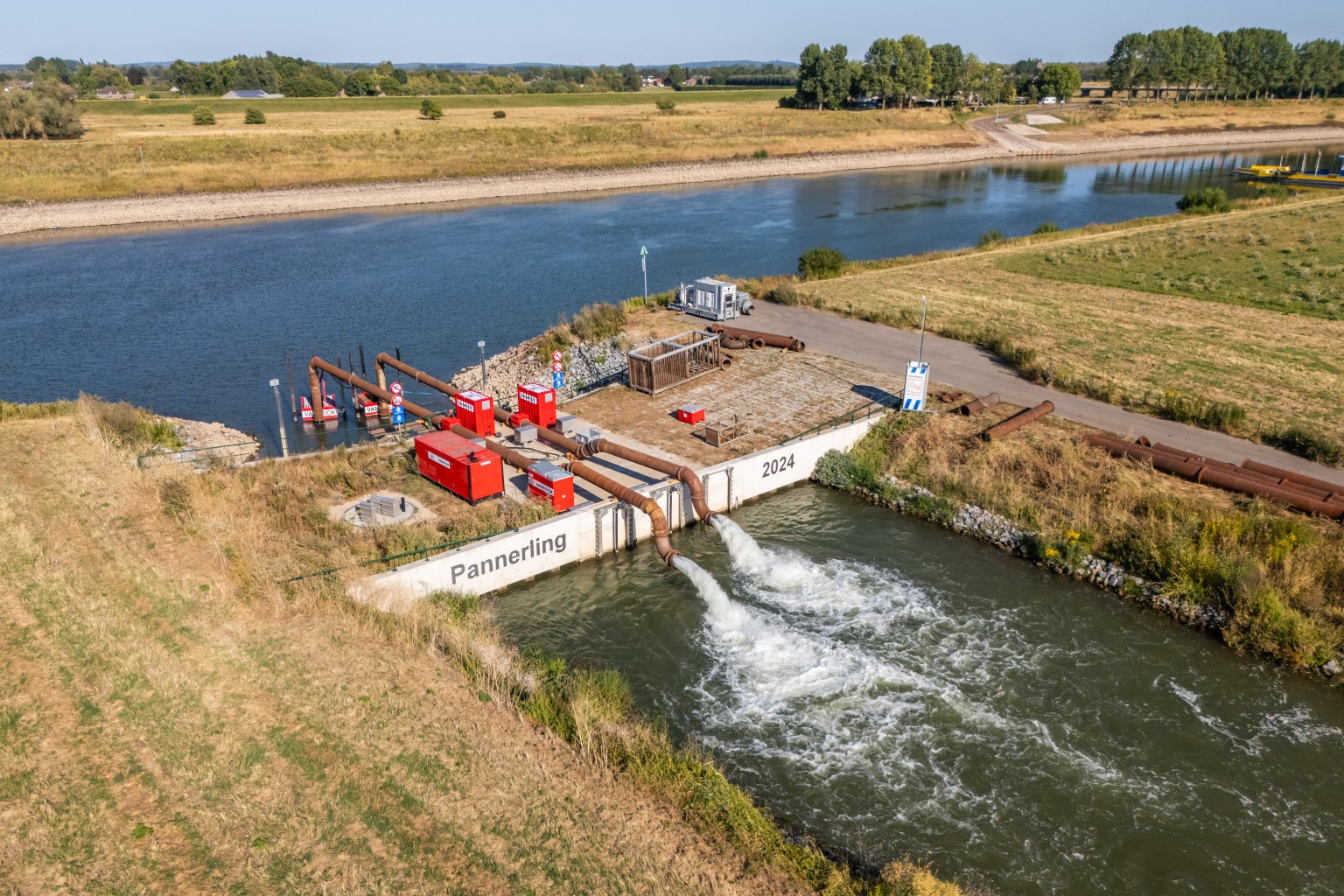 Noodpompen gemaal Pannerling Waterpompstation bij het kanaal Pannerling met rode apparatuur, leidingen en stromend water, omgeven door grasland en bomen.