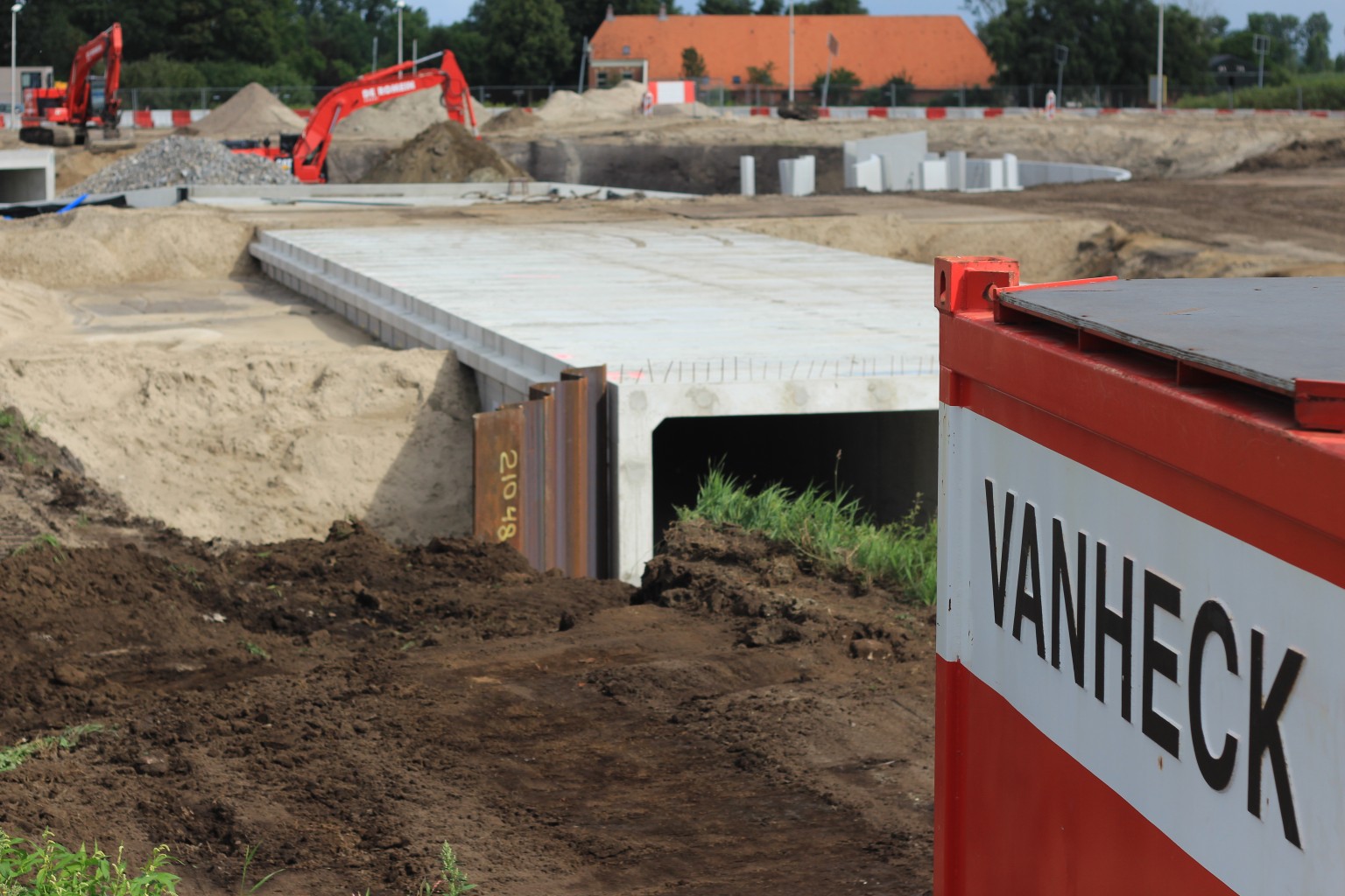 Rode container met opschrift "Van Heck" met op de achtergrond een nieuw gebouwde tunnel met daarachter twee graafmachines op een bouwplaats