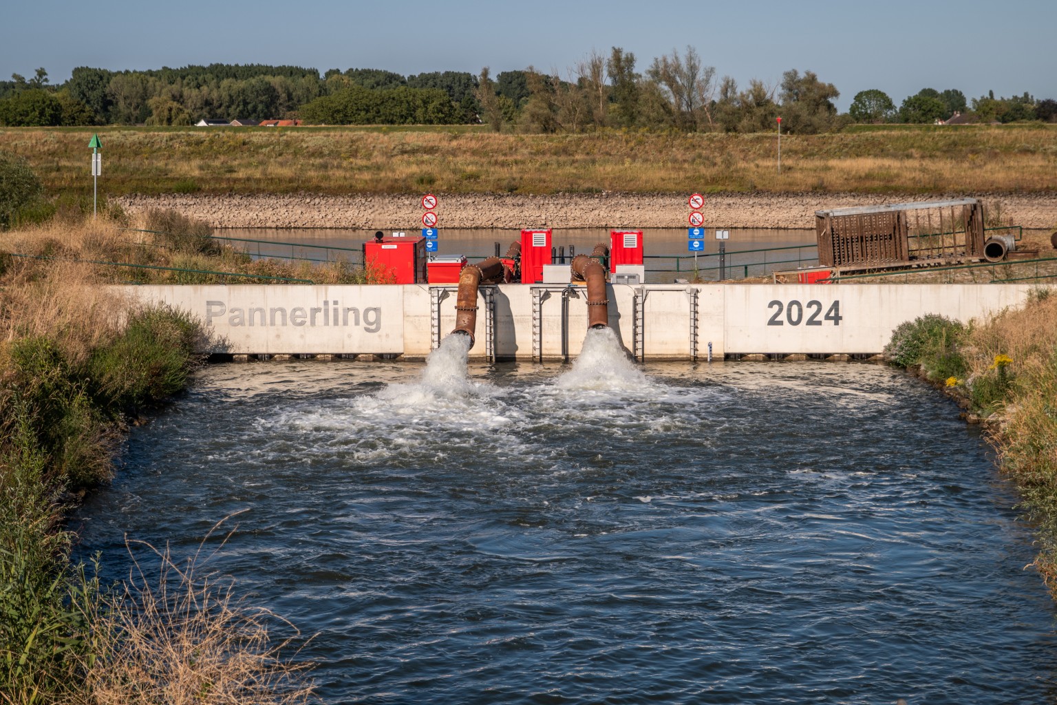 Gemaal Pannerling Water stroomt uit twee grote pijpen bij een constructie met de tekst "Pannerling 2024"; omringd door gras en bomen.