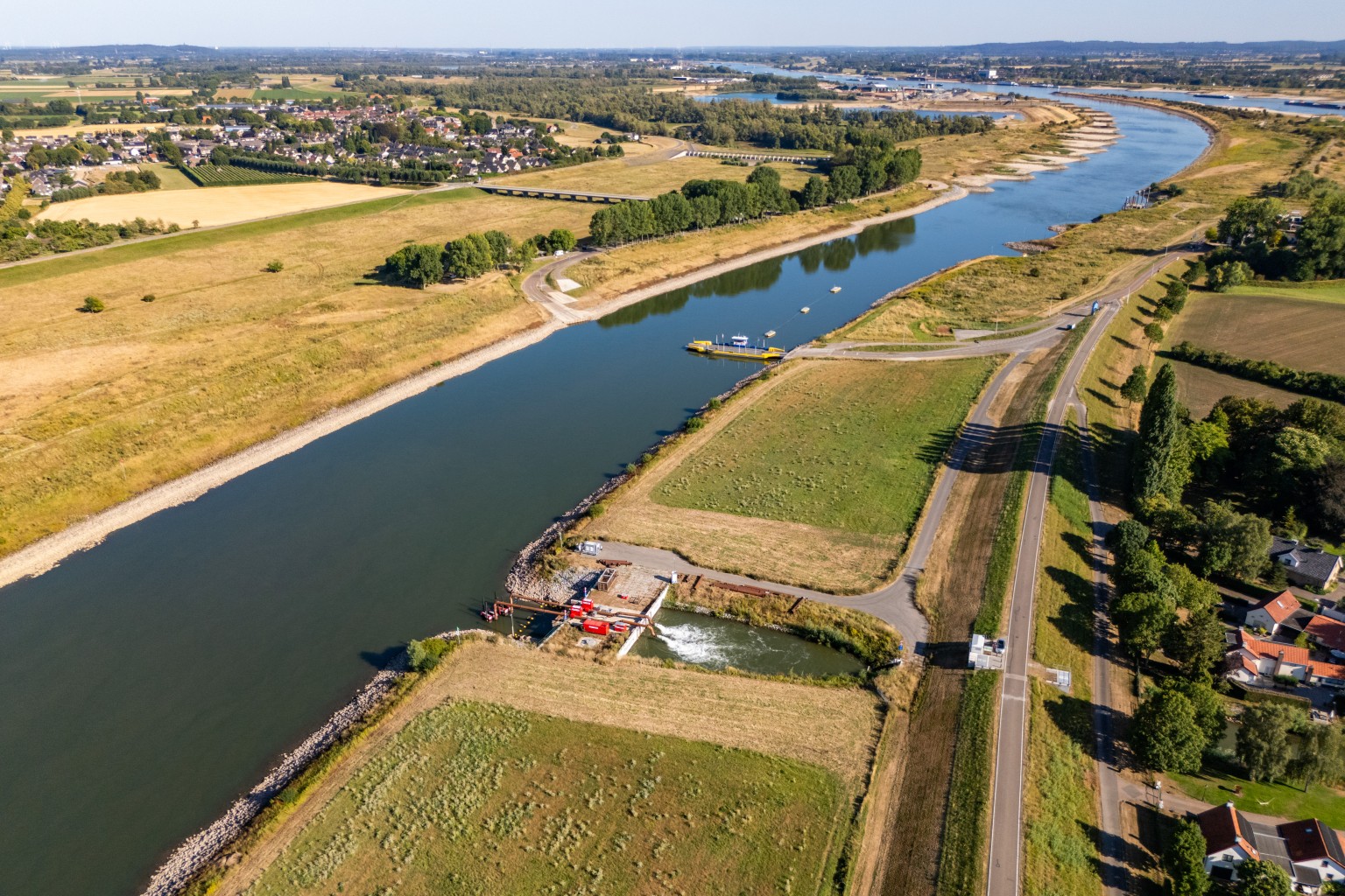 Pannerdensch Kanaal Luchtfoto van een kronkelende rivier met aangrenzende velden, een dijk en een klein dorp, onder een heldere blauwe hemel.