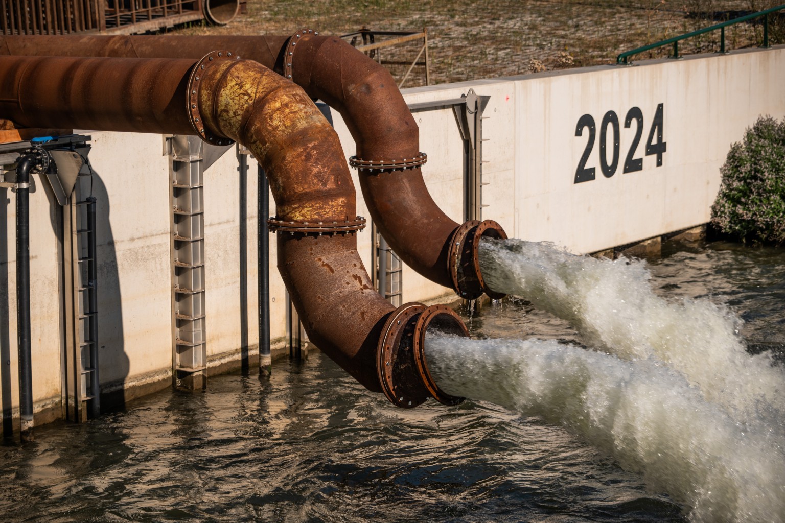 Uitstroom bij noodpompen Roestige waterleidingen spuwen water in een rivier. Op de achtergrond is een betonnen muur met het jaartal 2024 erop geschilderd.