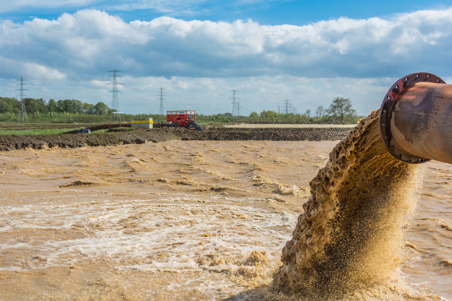Muddy water gushes from a large pipe into a dredging area, with fields and pumps in the background under a cloudy sky.