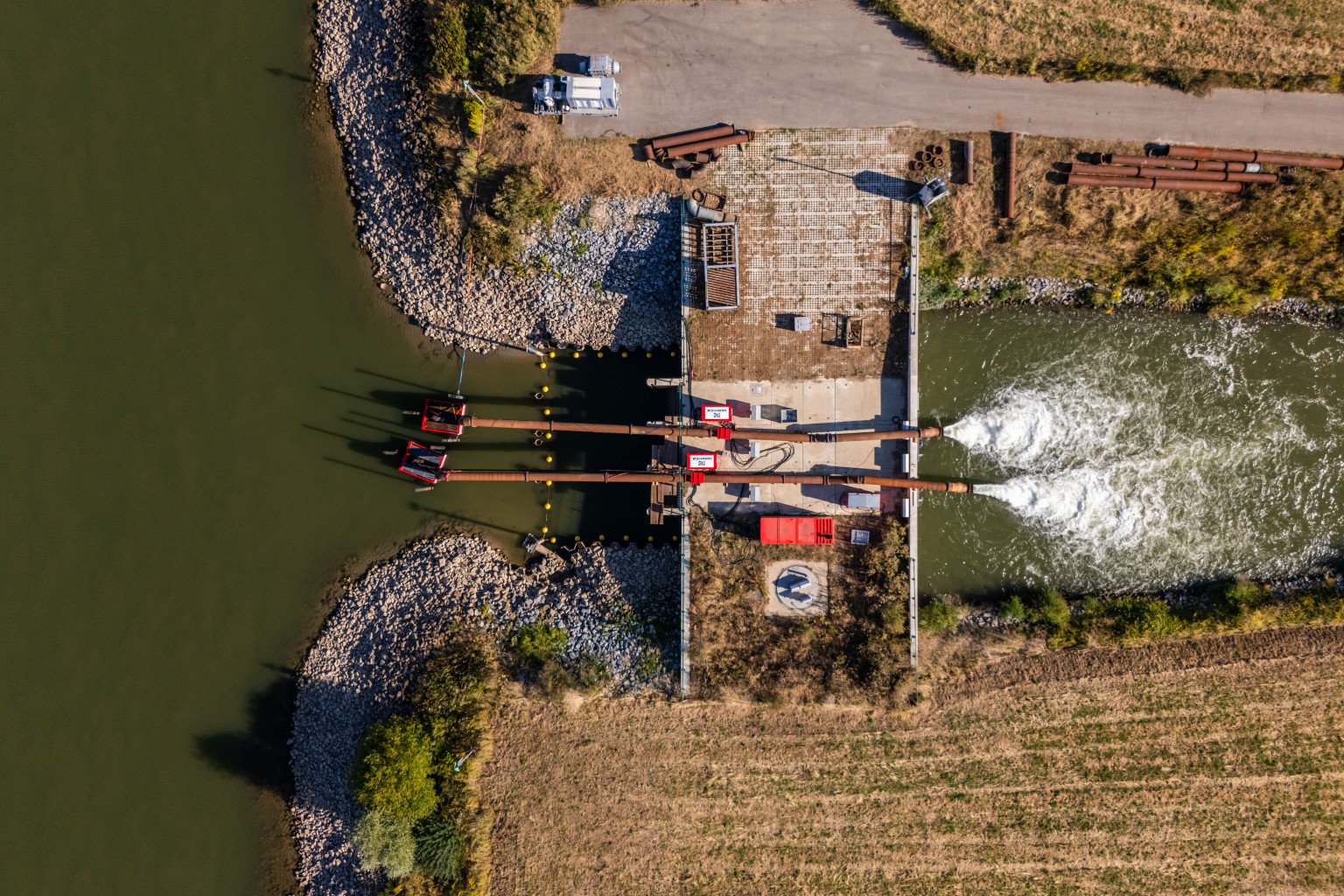 Bovenaanzicht gemaal Pannerling Luchtfoto van een gemaal met leidingen die water pompen in een kanaal, omgeven door groen en een geasfalteerd pad.