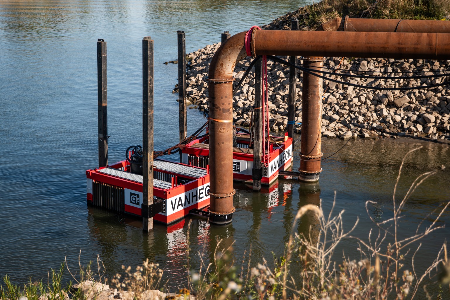 AFP600 bij Pannerling Drijvende waterpompinstallatie met "VAN HECK" opschrift in een rivier, omgeven door pijpleidingen en rotsachtige oever.