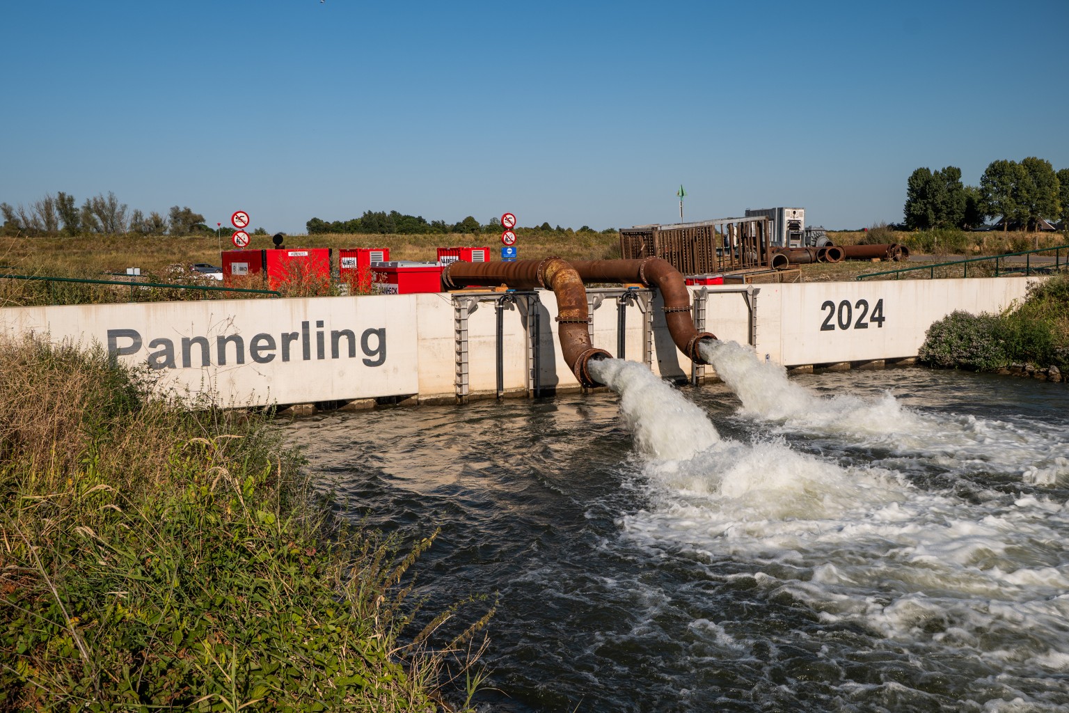 Pompen bij Gemaal Pannerling Water stroomt uit twee grote pijpen bij een constructie met de tekst "Pannerling 2024"; omringd door gras en bomen.