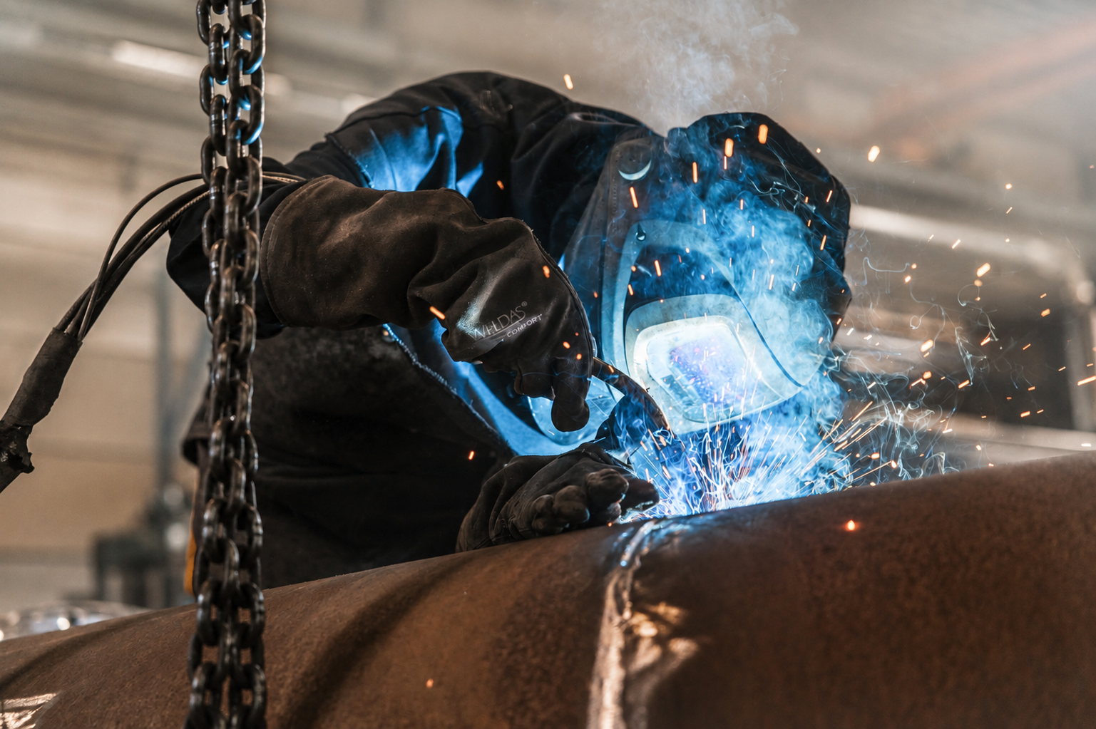 Lasser bezig met bocten Persoon met lashelm en handschoenen last metalen buis, omgeven door blauwe lichtflitsen en vonken in een industriële omgeving.