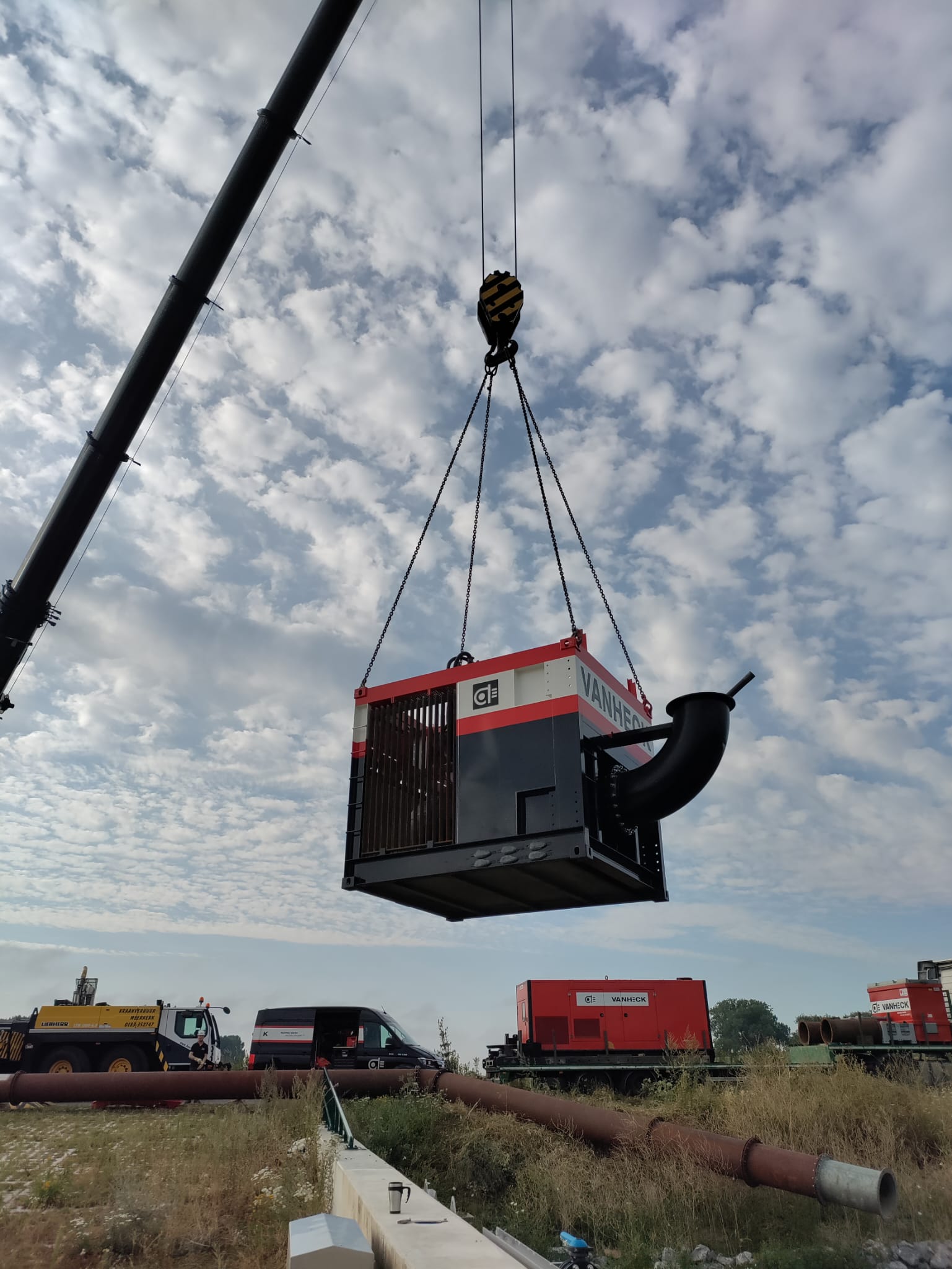 Inhijsen van de tijdelijke pomp Een kraan tilt een grote machine in een bouwgebied tegen een bewolkte hemel. Op de achtergrond staan voertuigen en pijpen.