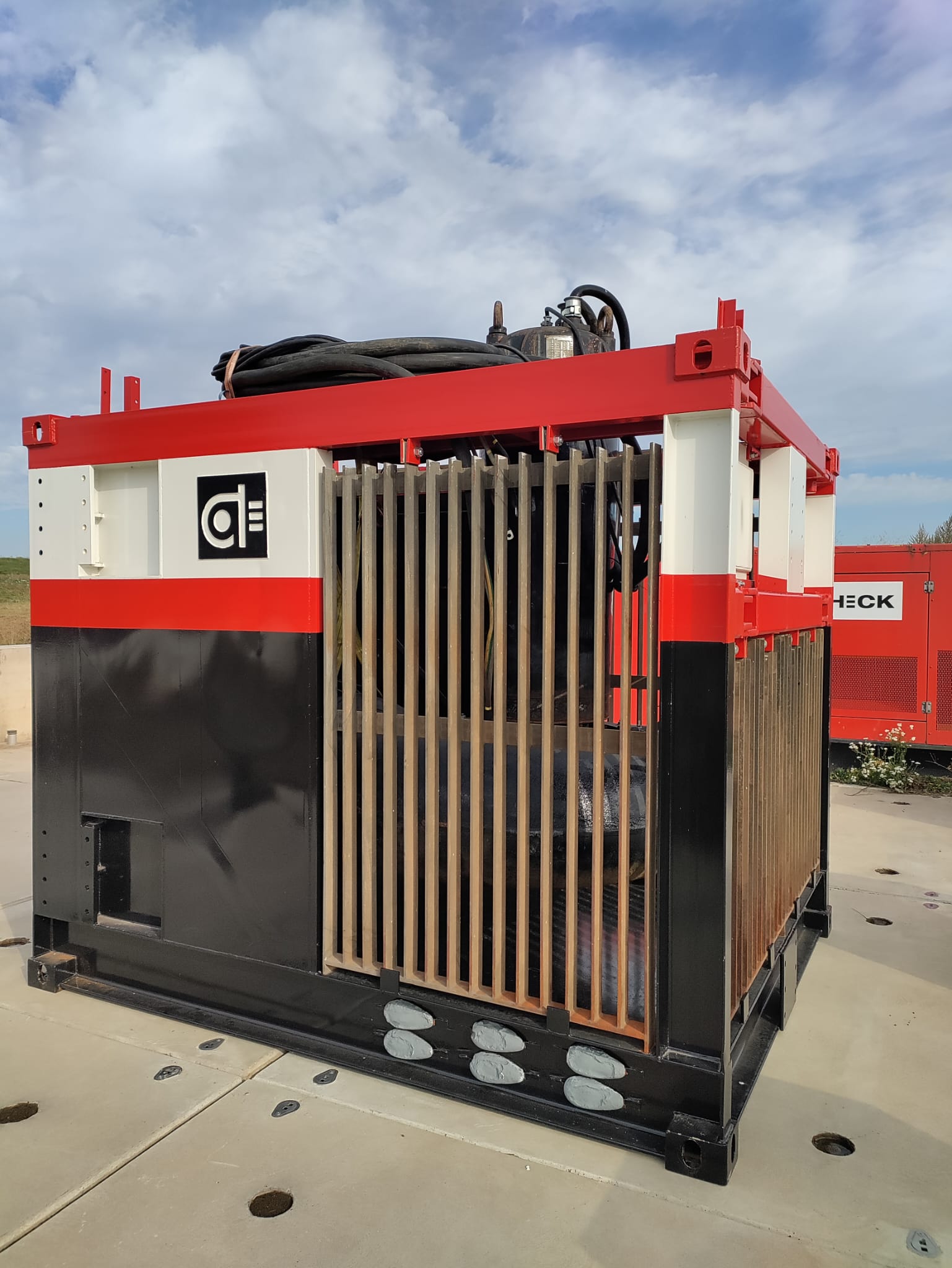 Afp600 klaar voor plaatsing Industriële machine met rode en witte accenten, omgeven door metalen tralies, geplaatst op een betonnen vloer onder een bewolkte hemel.
