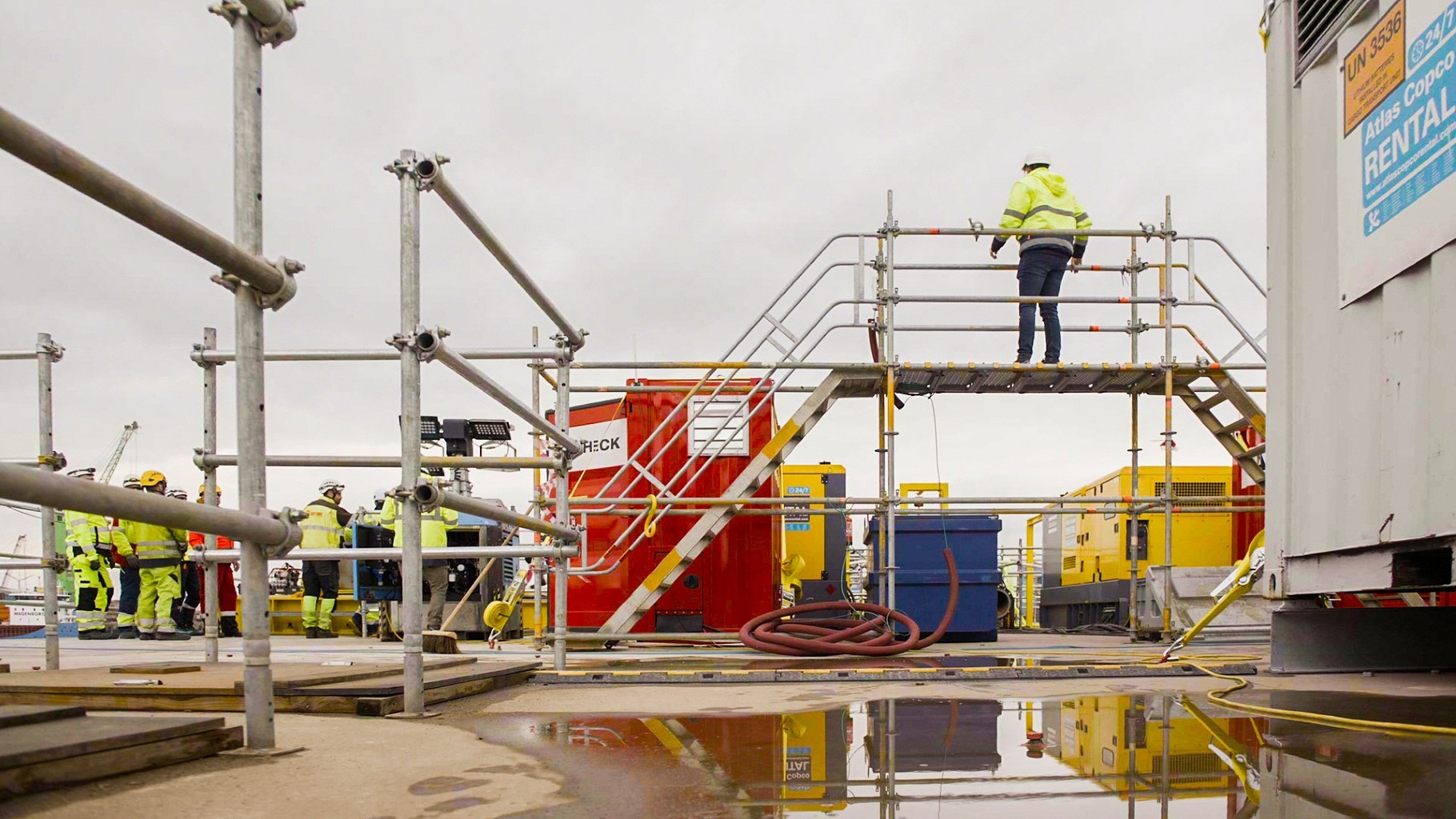 Bouwplaats met steigers en arbeiders in veiligheidskleding. Een persoon staat bovenop een platform. Er liggen plassen water op de grond.