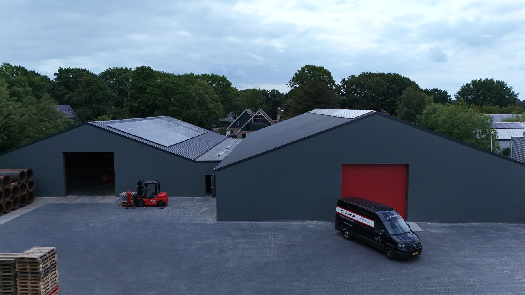 Grijs magazijngebouw met rode deuren, parkeerplaats met een zwarte bestelwagen en een heftruck, omringd door bomen.
