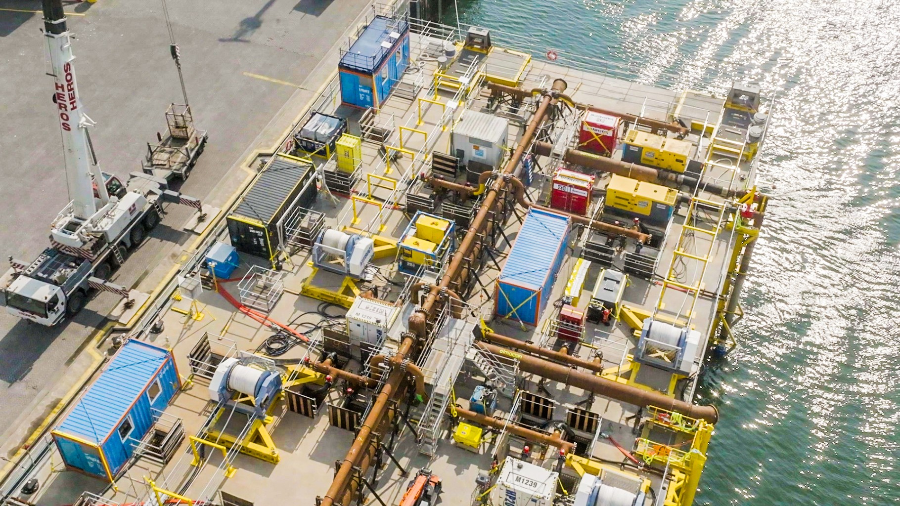 Luchtfoto van een industriële installatie aan de waterkant met containers, kabels en machines, naast een kraan op de kade.
