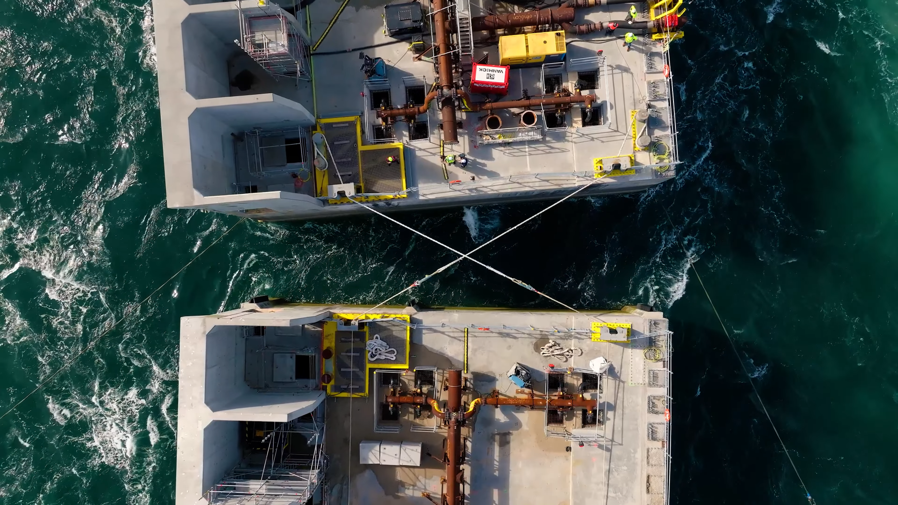 Luchtfoto van twee grote betonnen platforms op zee, verbonden door metalen kabels boven golvend water. Mensen staan op het bovenste platform.