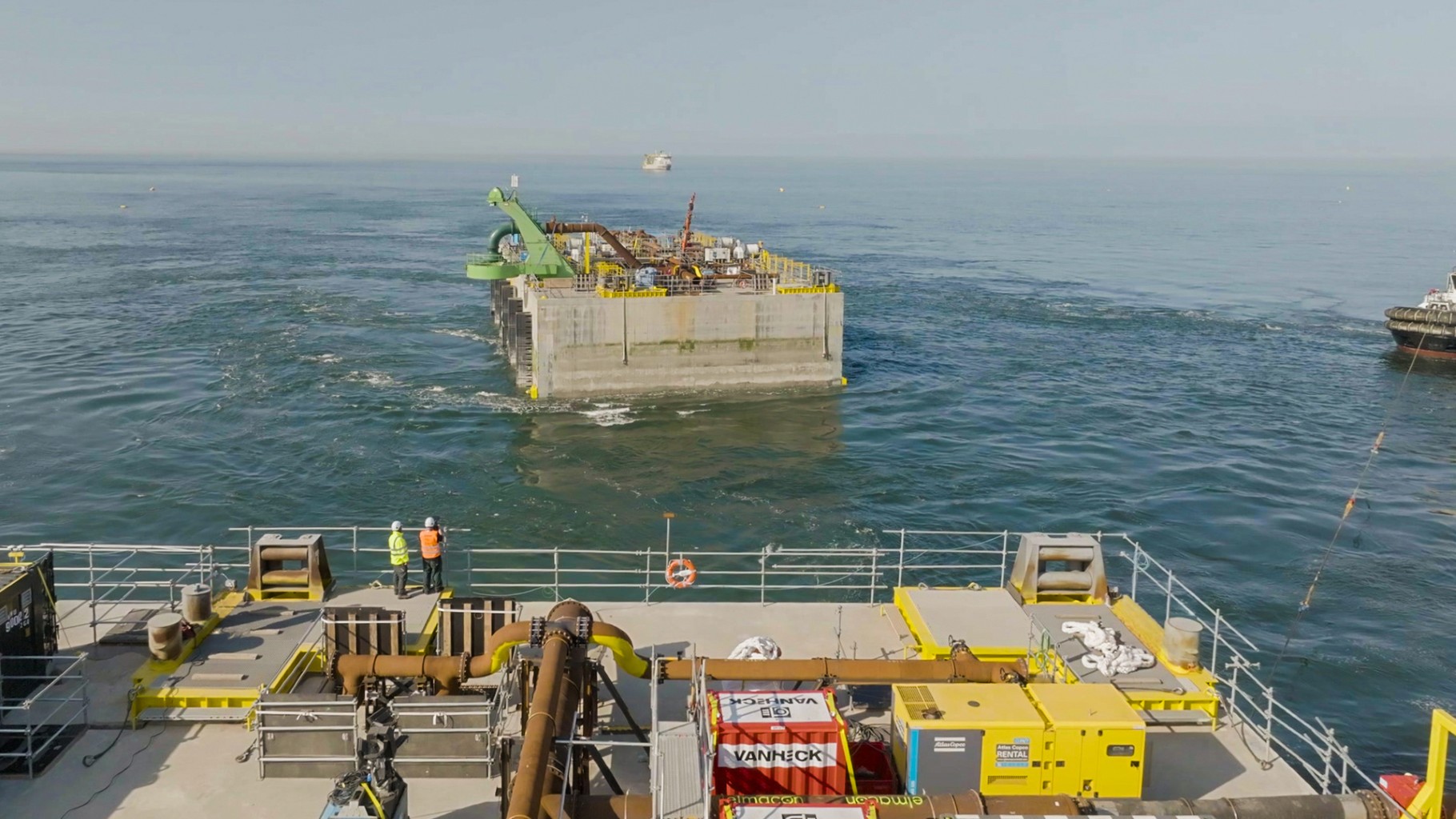 Groot betonnen platform in zee, omringd door water. Twee mensen staan op de voorgrond op een schip met gele apparatuur.