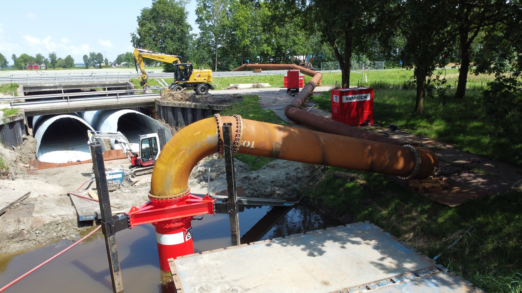 Rode waterpomp op pootjes staat in een watergang met een stuk leidingwerk voor een bouwput met twee stalen kokers onder een weg door in een groene omgeving.