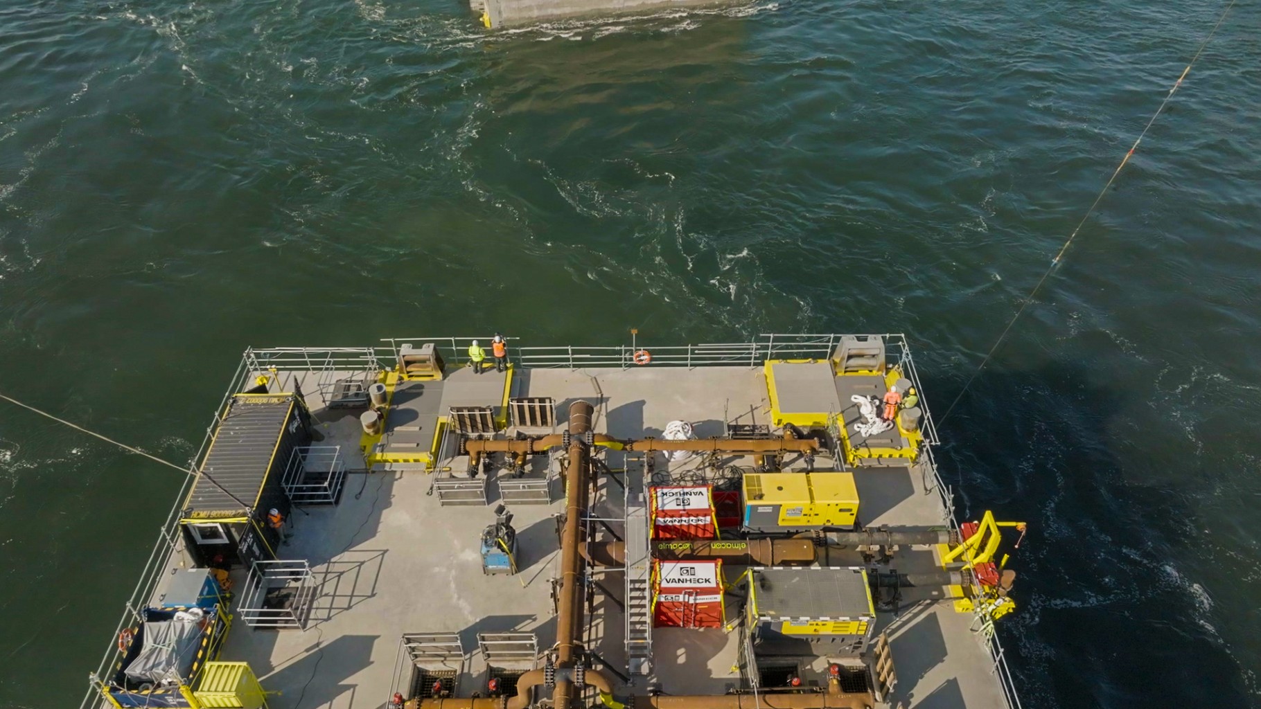 Luchtfoto van een groot caisson op zee, met diverse apparatuur en bemanning aan dek, omringd door diepblauw, golvend water.