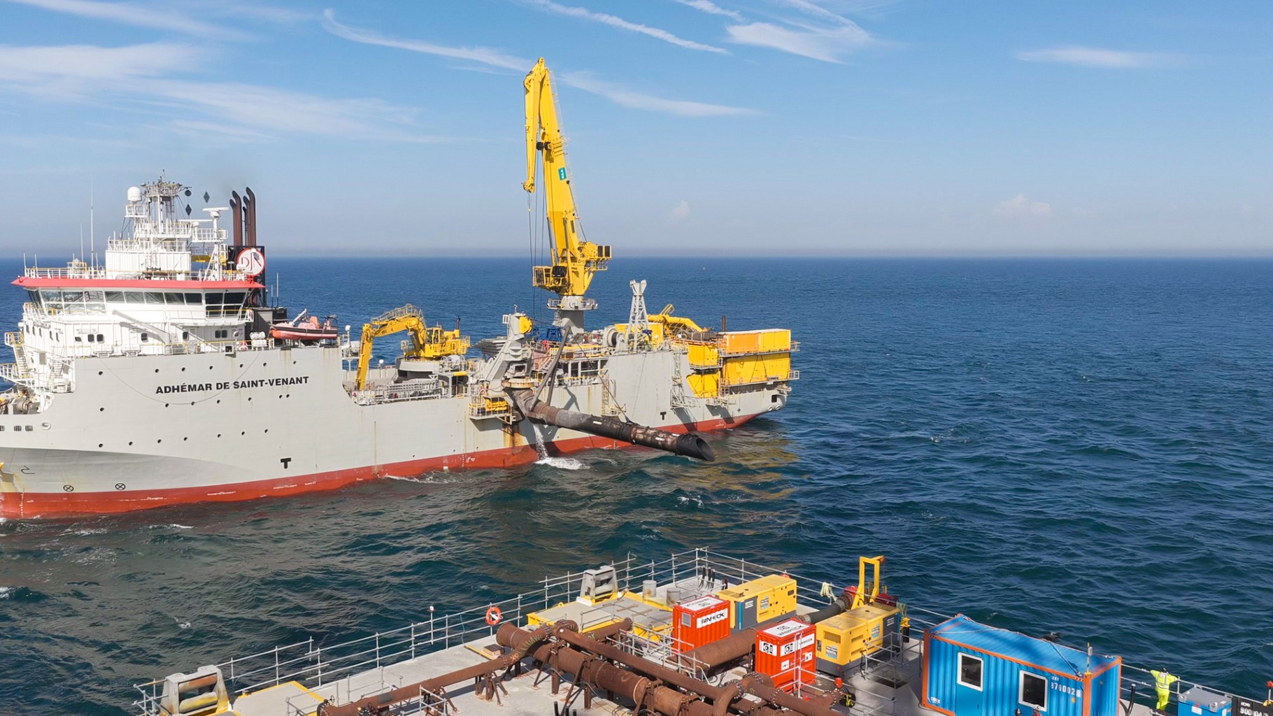 Grote schip met kraan, gele en rode containers aan dek, opereert op de oceaan. Zonnige dag met blauwe lucht en kalme zee.