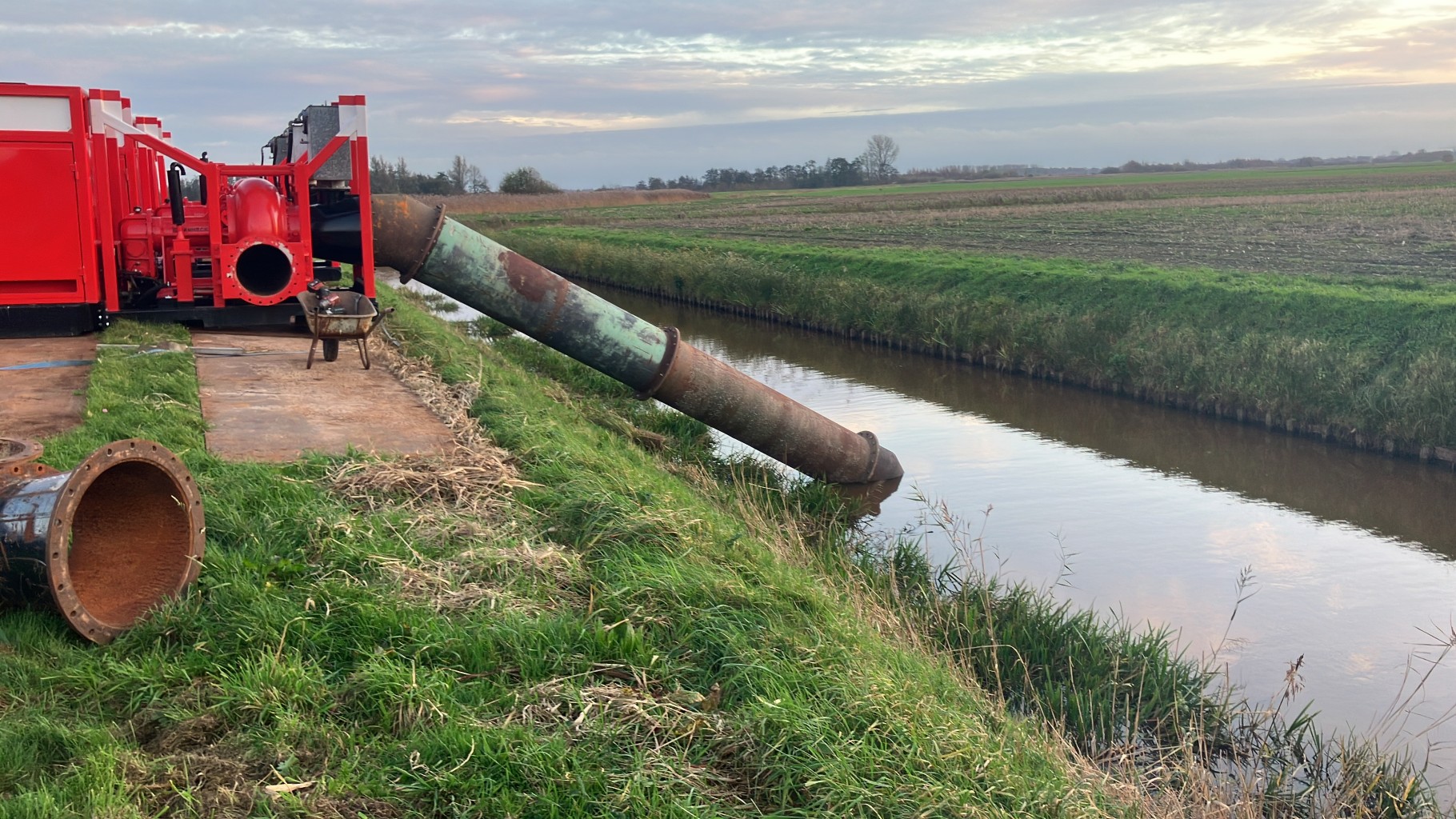 Een rode pomp staat op een kade met een stuk leidingwerk dat in een sloot staat midden in de omgeving van een weiland