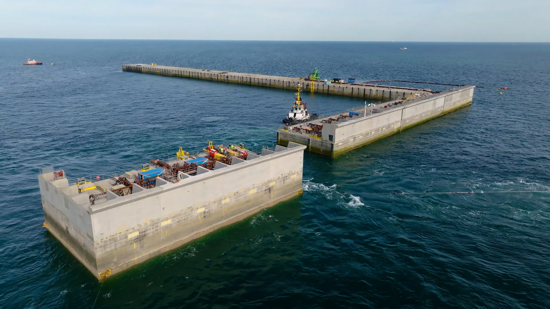 Luchtfoto van een kunstmatig eiland in aanbouw met schepen op de achtergrond. Grote betonnen structuren drijven in het water, begeleid door een sleepboot.