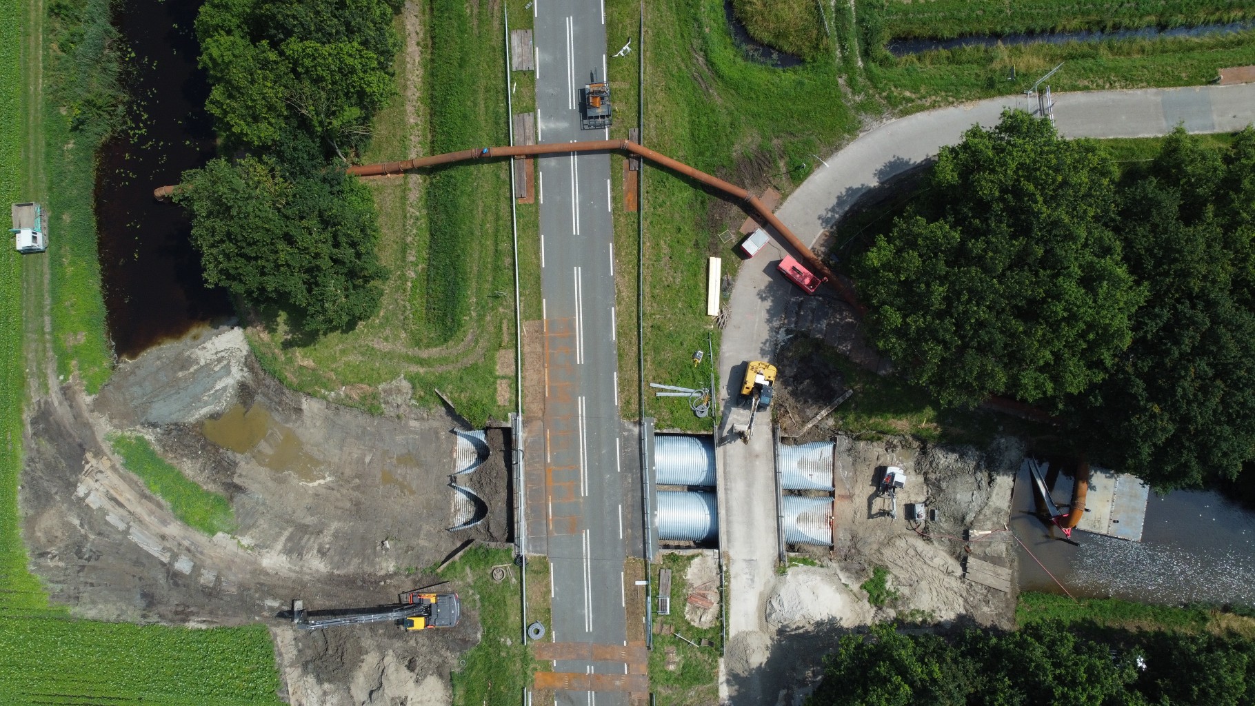 Luchtfoto van een weg in een groene omgeving met twee duikers onder een weg door nabij een bouwput. een stalen leiding loopt over de weg heen