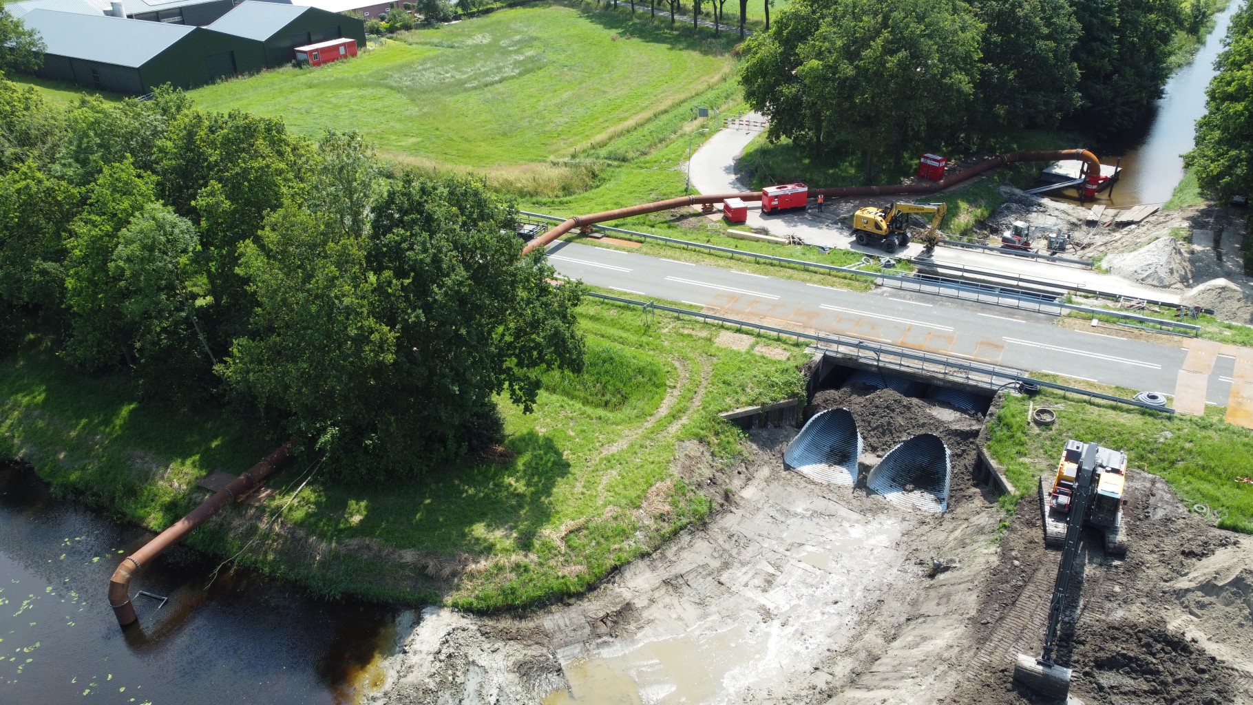 Bouwplaats in groene omgeving met een graafmachine en een stalen leiding die tussen twee sloten loopt en een weg kruist.