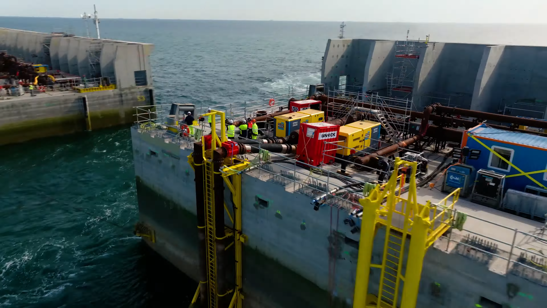 Luchtfoto van een bouwplaats op zee met grote betonnen structuren, machines en werkers in veiligheidskleding langs de kustlijn.