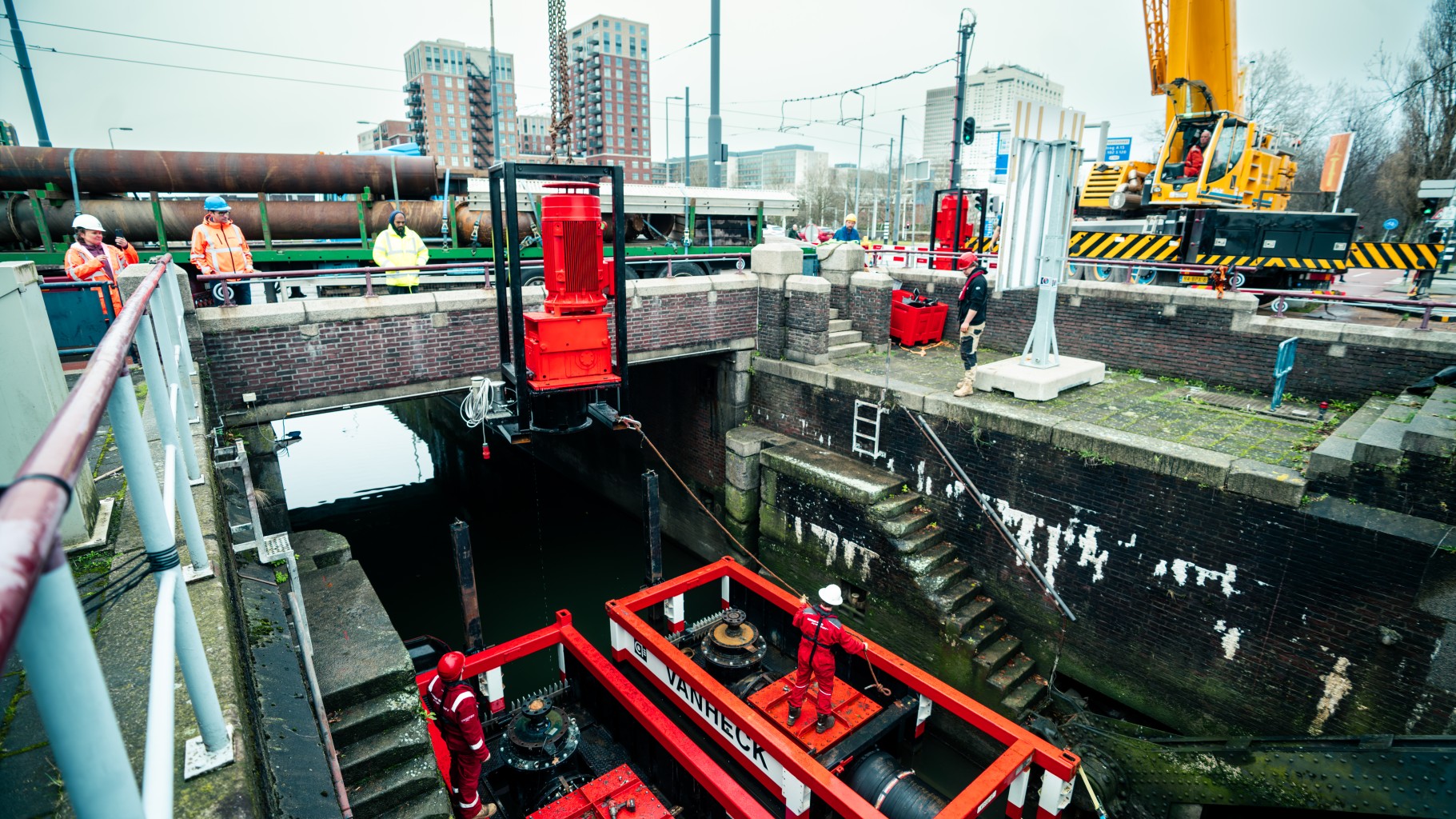 Demobilisatie tijdelijke pompset Een elektromotor wordt gehesen uit een sluis met daarin twee pompsets met daarop een monteur. De pompen staan in een sluis in een stadse omgeving