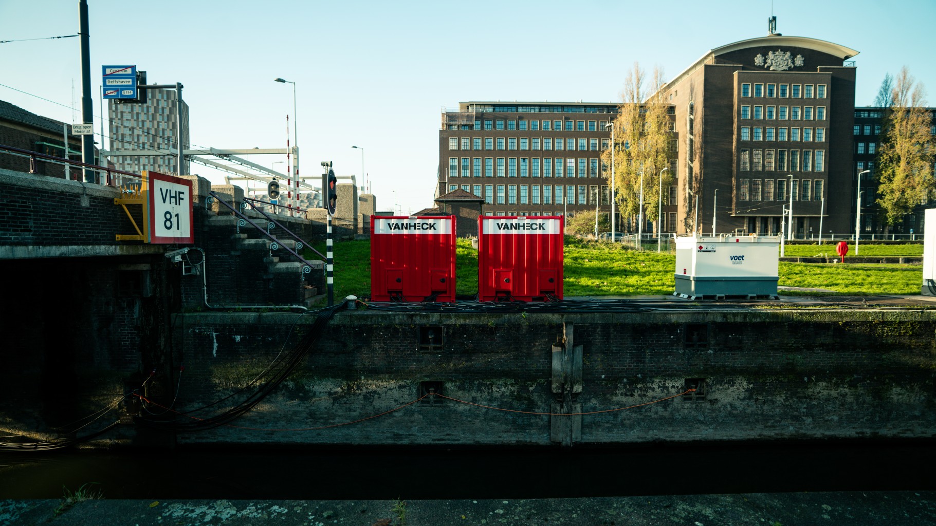 Twee rode containers met opschrift "van Heck" staan op een kade muur nabij een brug en groot gebouw