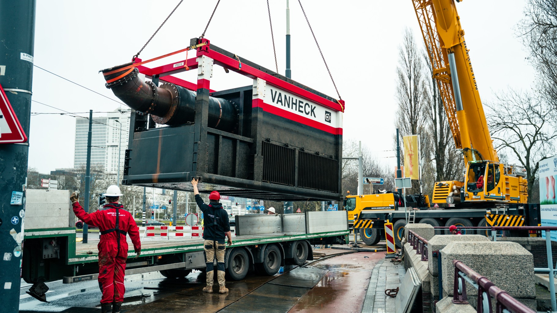 Afbouw VP800 Grote zwart-rood met witte pompcontainer wordt door een gele kraan geladen op een trailer midden op straat. twee monteurs begeleiden het hijsen