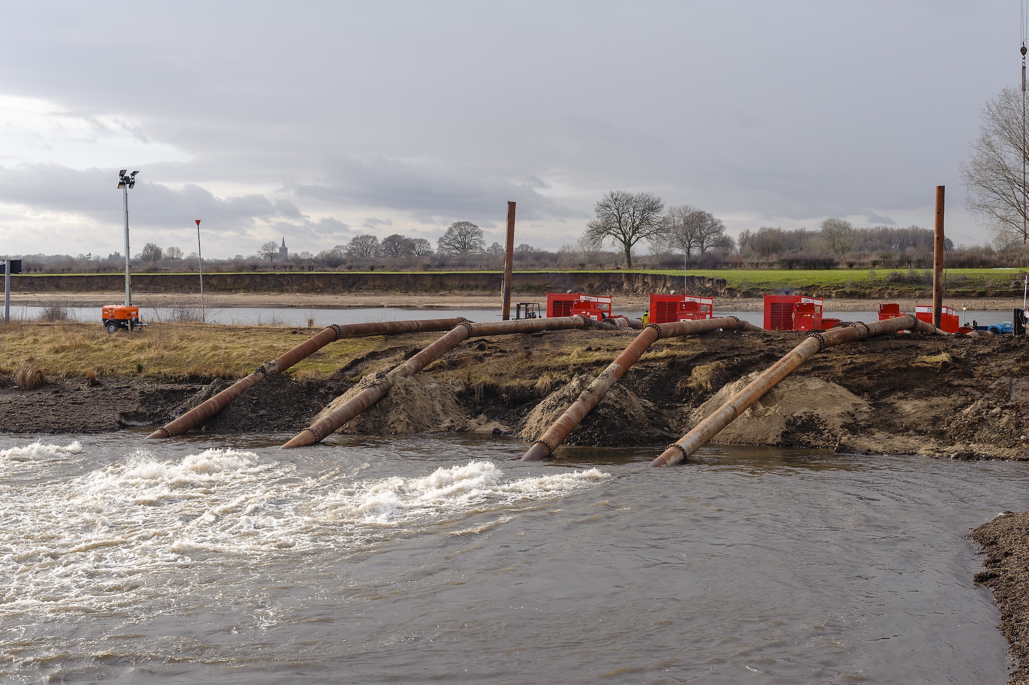 Large pipes and red waterpumps near a riverbank with turbulent water, under a cloudy sky and surrounded by grassy landscape.