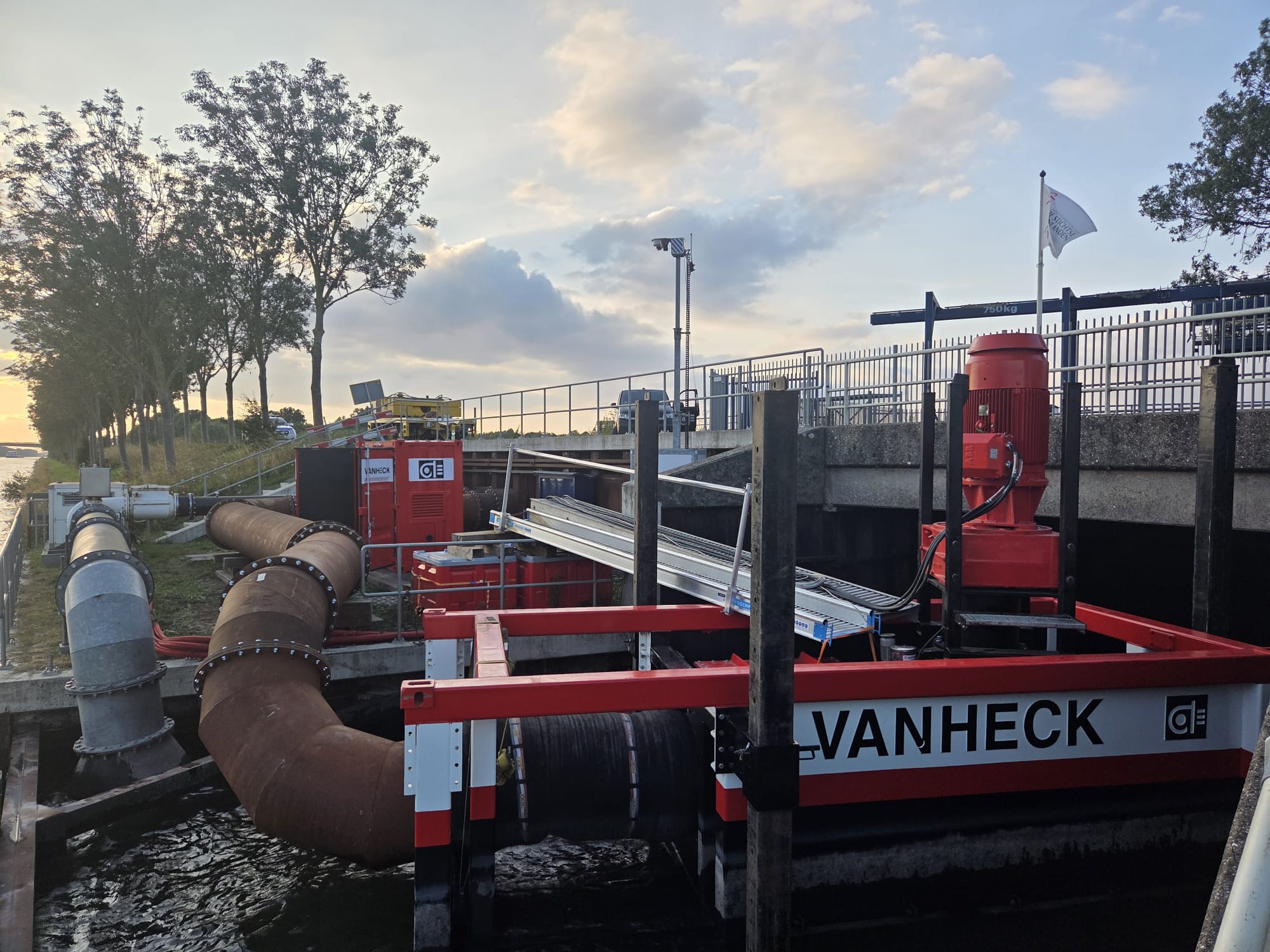 Pomp met elektromotor en tekst "Van Heck" met leidingwerk naast een brug. Links een bomenrij onder een licht bewolkte lucht in de avond.