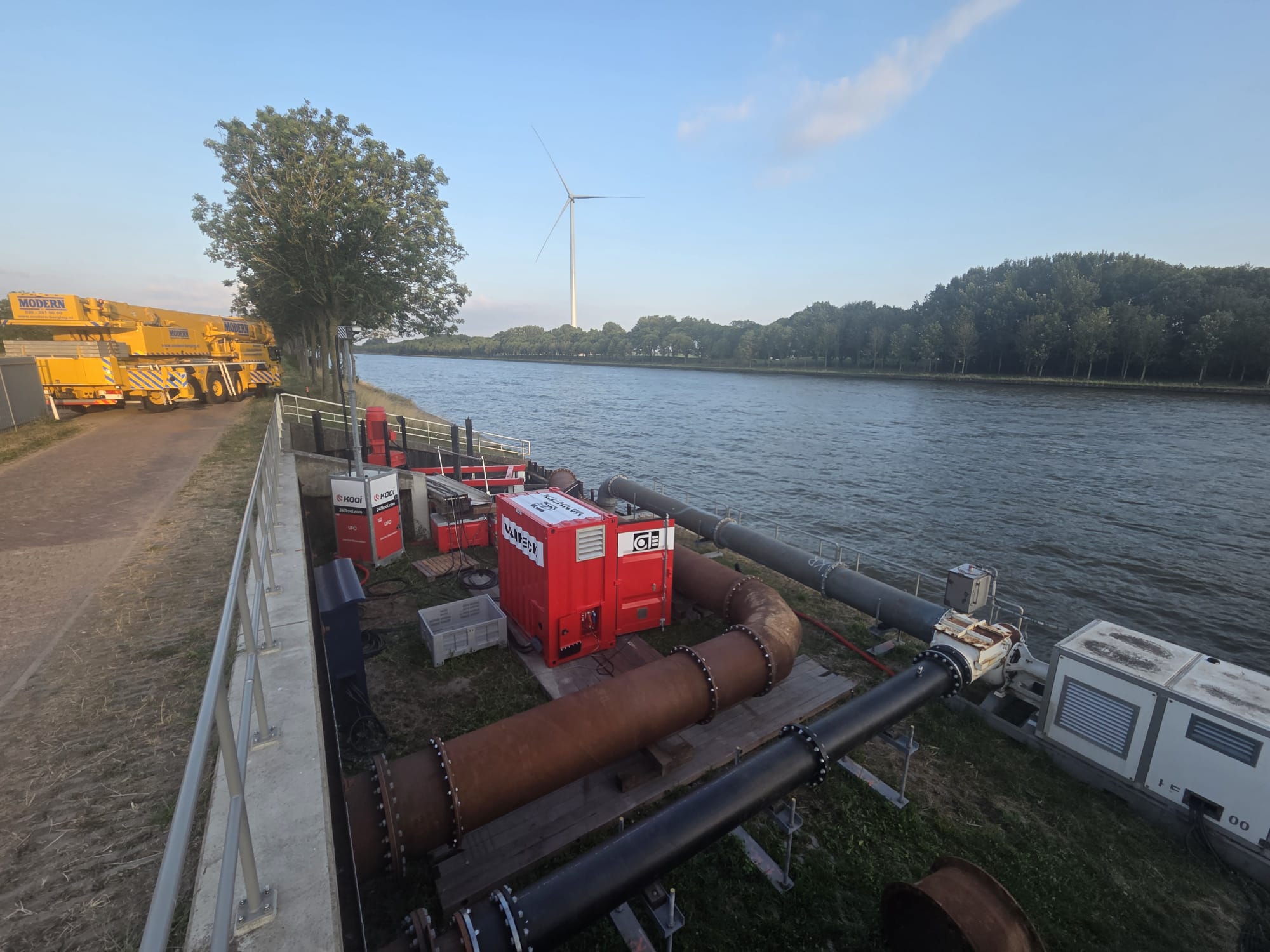 Technisch bouwmateriaal langs een kanaal met aan de rechterzijde een bomenrij onder een blauwe lucht
