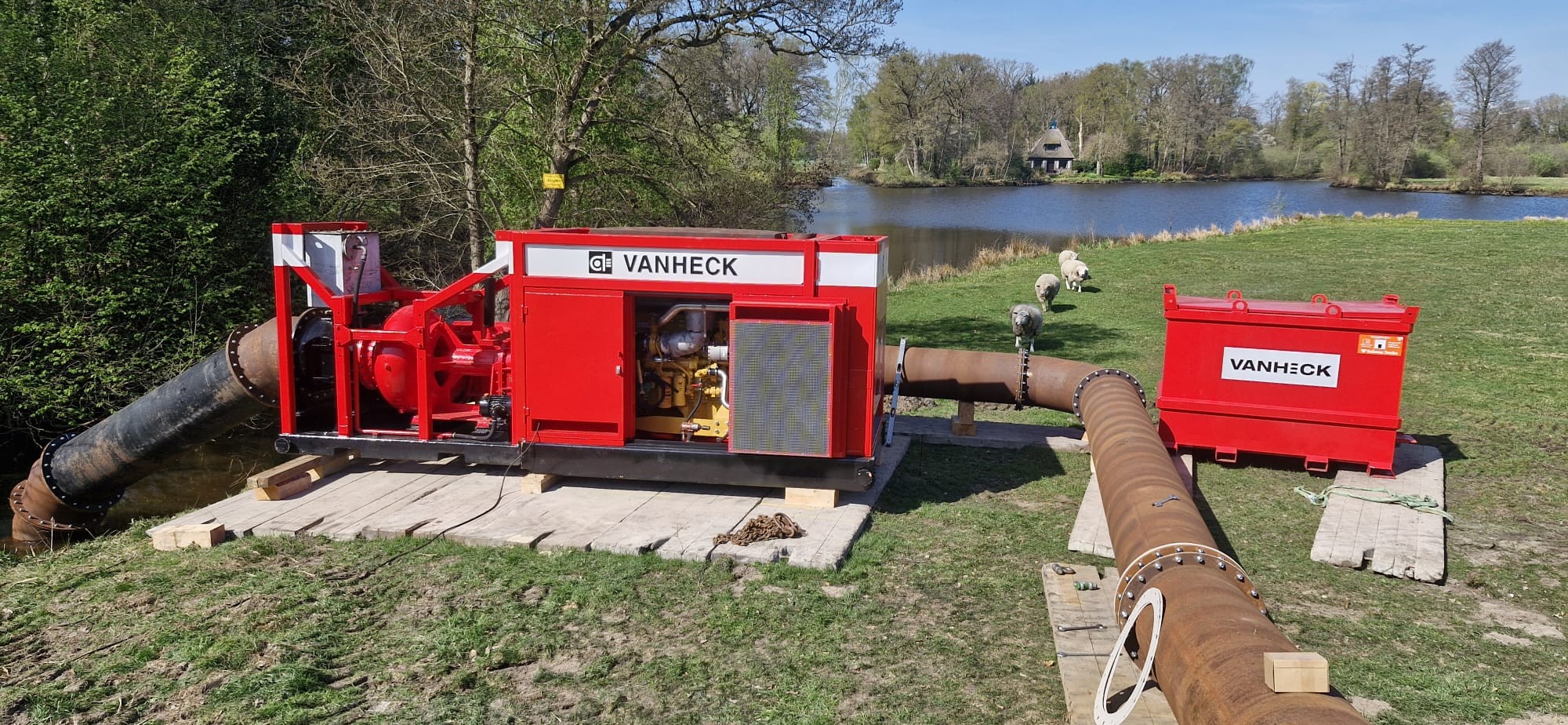 Tijdelijke pompinstallatie HK500 Rode pomp met opschrift "Van Heck" met een leiding in het water op een platform van planken op een weiland nabij een kanaal en boom met op de achtergrond schapen