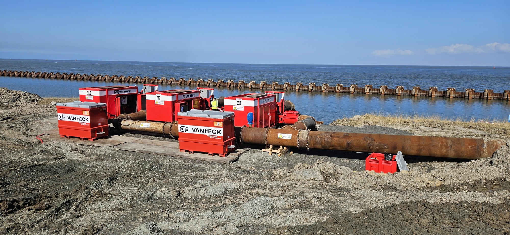 drie rode waterpompen en twee rode tanks voor een stuk leidingwerk in een ruige zandomgeving nabij de zee met een stuk damwand