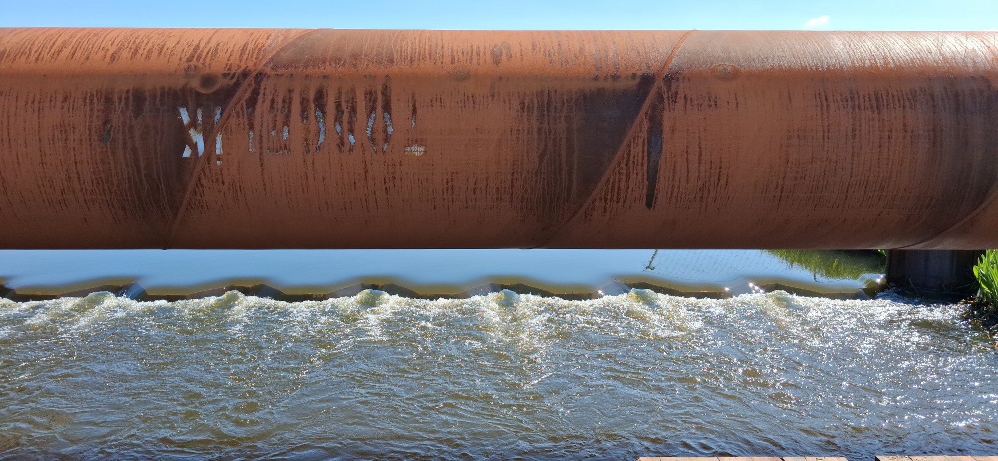 Leidingwerk over waterstroom Een stalen leiding met oxidatie ligt over een stroom met water dat stroomt en wordt gebroken door een damwand