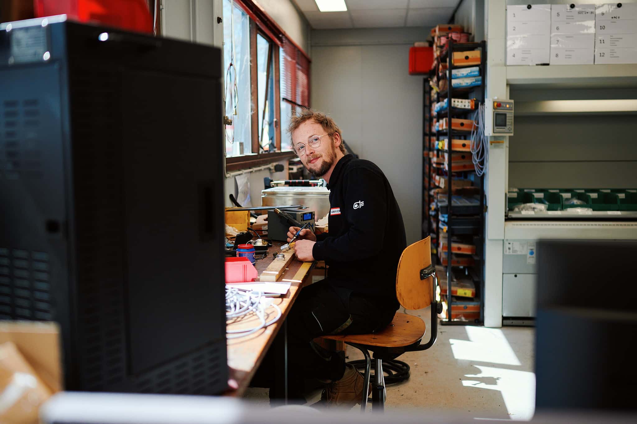 Engineering Man zit aan bureau in werkplaats met elektronica en gereedschap, kijkend naar de camera. Achtergrond toont planken met dozen en kabels.