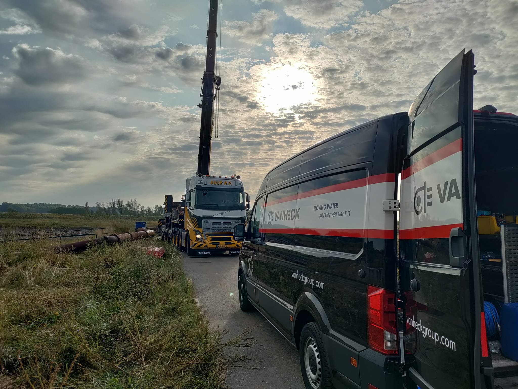 Opbouw noodpomp bij droogte Zwarte bestelwagen van Van Heck en grote kraanwagen op een weg, omgeven door grasland en bewolkte hemel met zonnestralen.