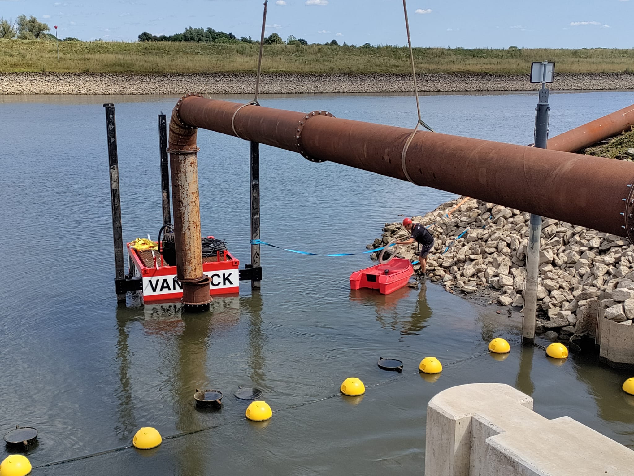 Tijdelijke pomp bij installatie Persoon werkt aan een waterbouwproject, staand op stenen bij water en een grote pijpleiding; boei-afzetting aan de voorgrond.