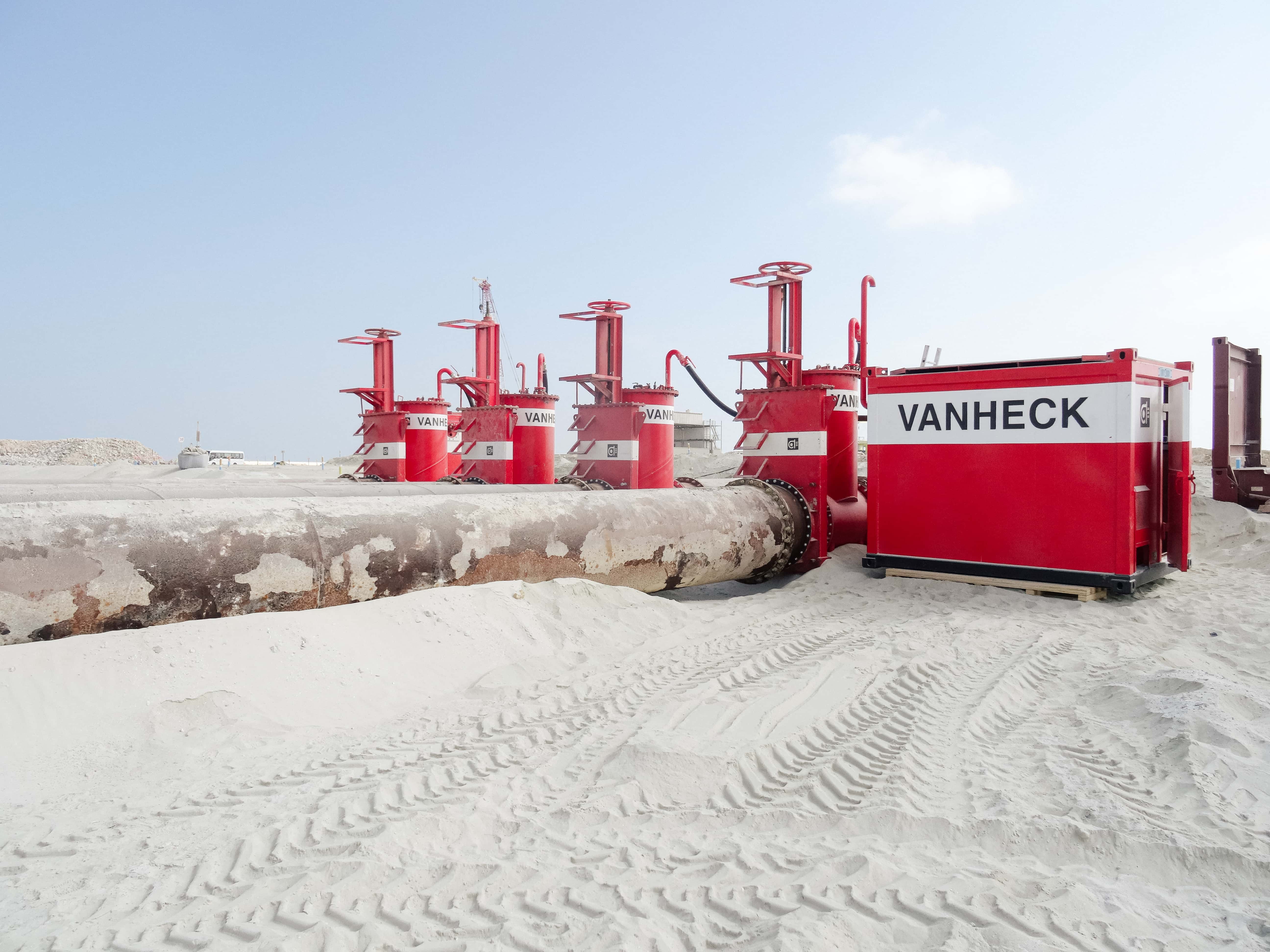 Rode hevel apparatuur en leidingen op een zandig terrein, met de naam "VANHECK" zichtbaar op een container. Lichte wolken aan de hemel.