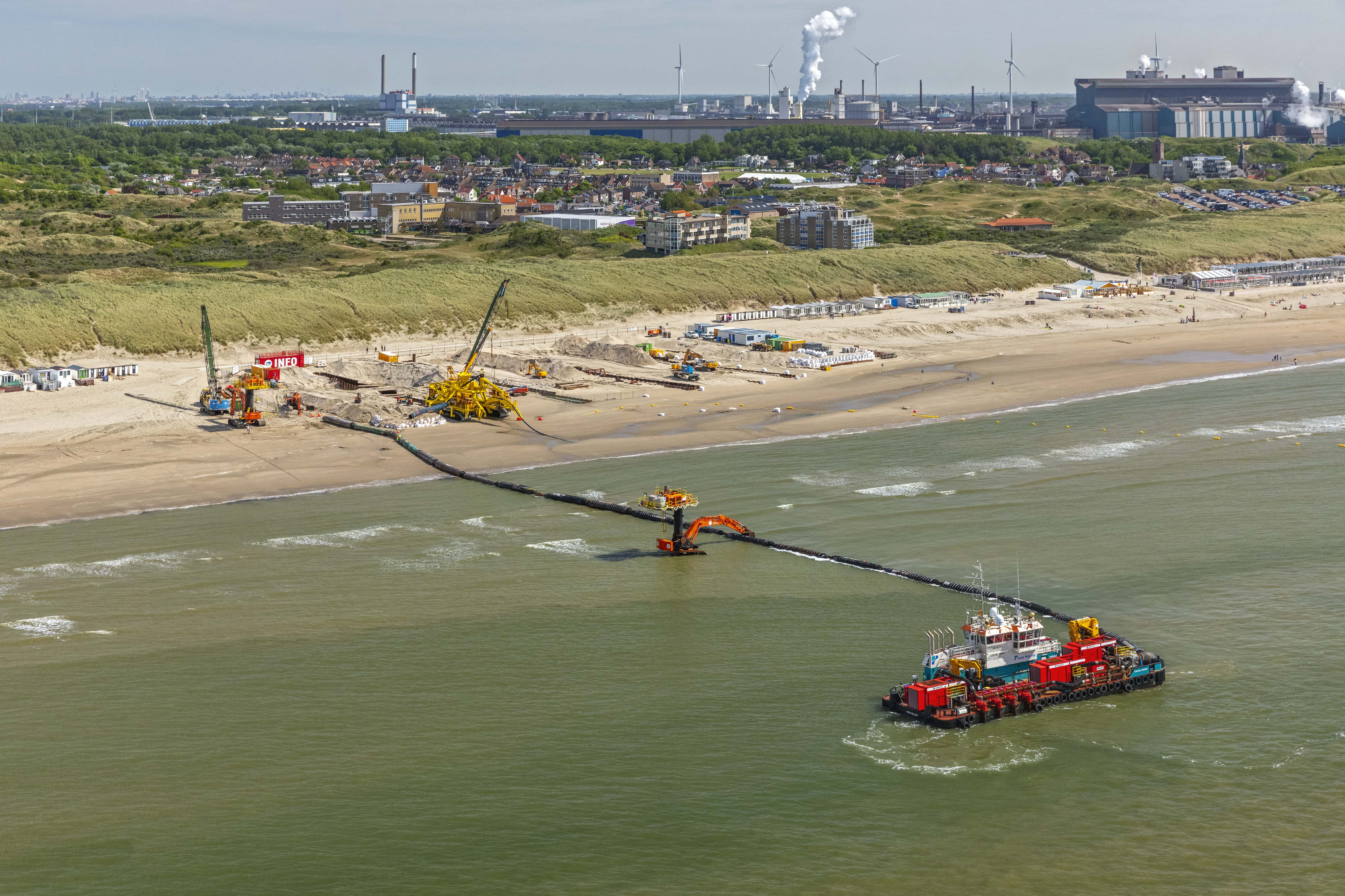 Kabeltrencher IJmuiden Multicat uitgerust met waterpompen die de kabeltrencher op het strand voorziet van water. Het strand is vol met bouwmachines en op de achtergrond is industrie te zien