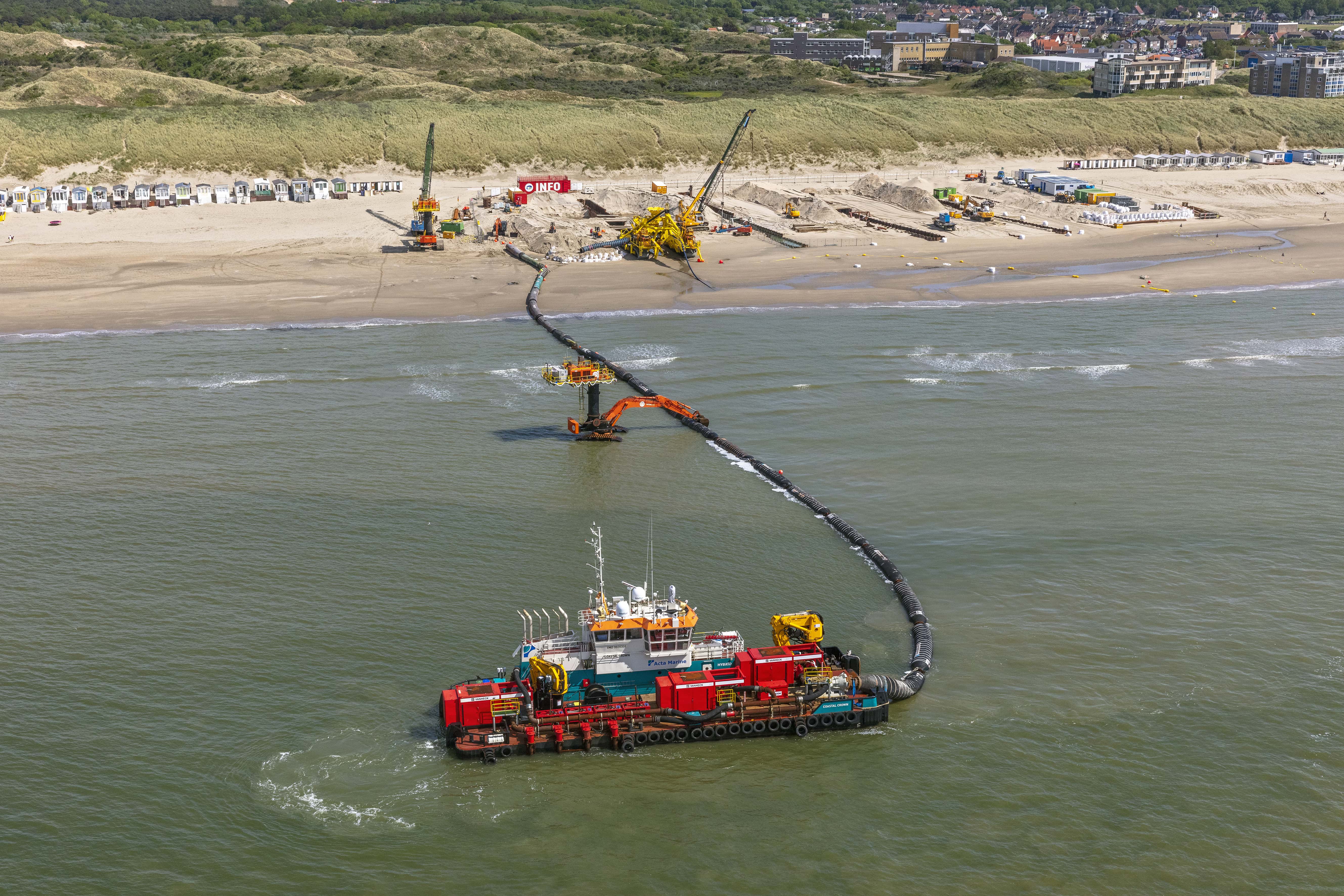 Van Heck bij kabelleggen Barge equipped with red waterpumps labeled with "Van Heck". The waterpumps supply water to the yellow cabletrencher on the beach. The background contains a dune landscape with houses. The beach is full of equipment. the sea is calm