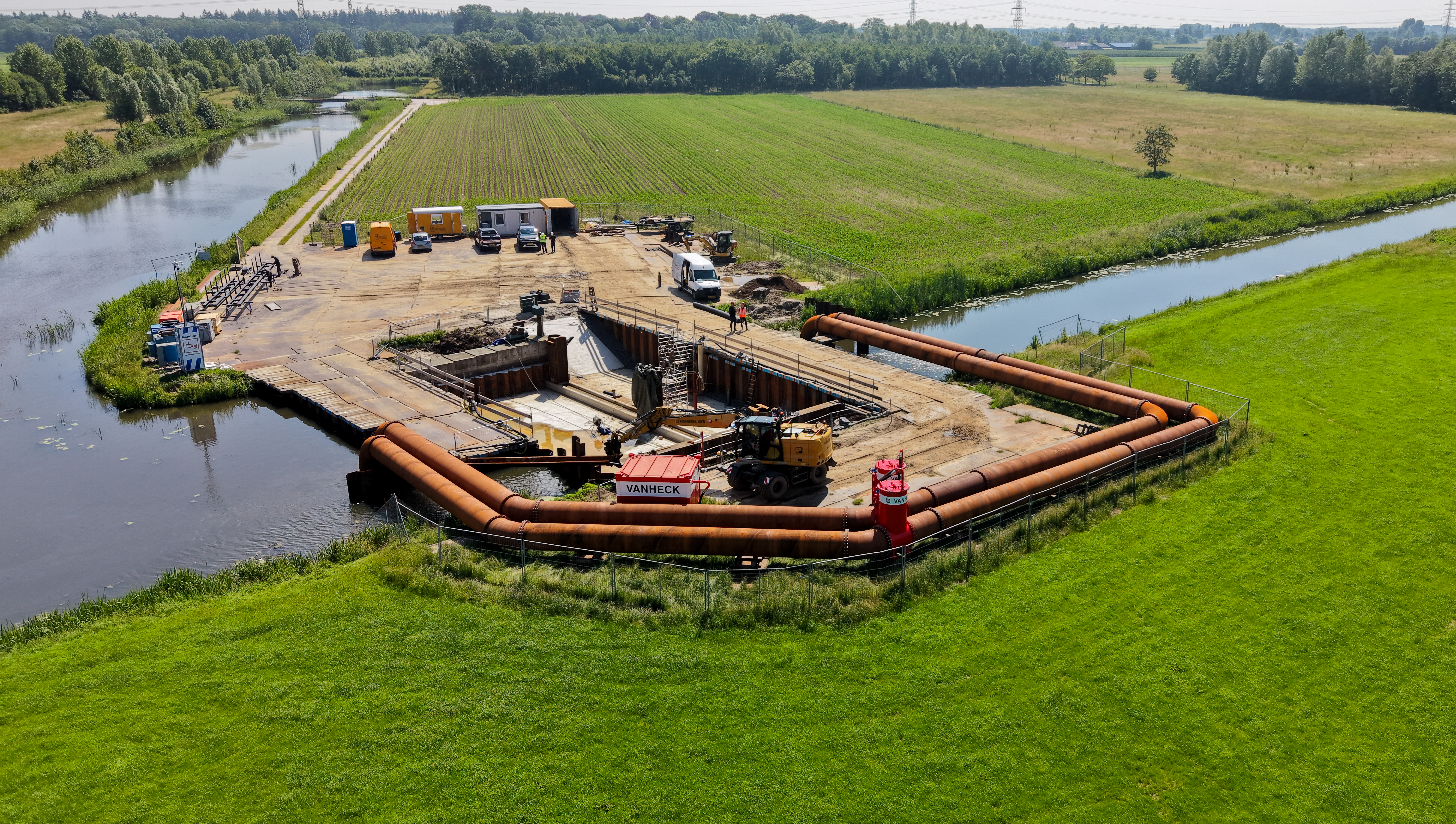 Overzichtsfoto van een dubbele hevel Twee stalen leidingen met rood materieel nabij een bouwkuip op een bouwplaats midden in een groen landschap van landerijen en bomen en een kanaal.