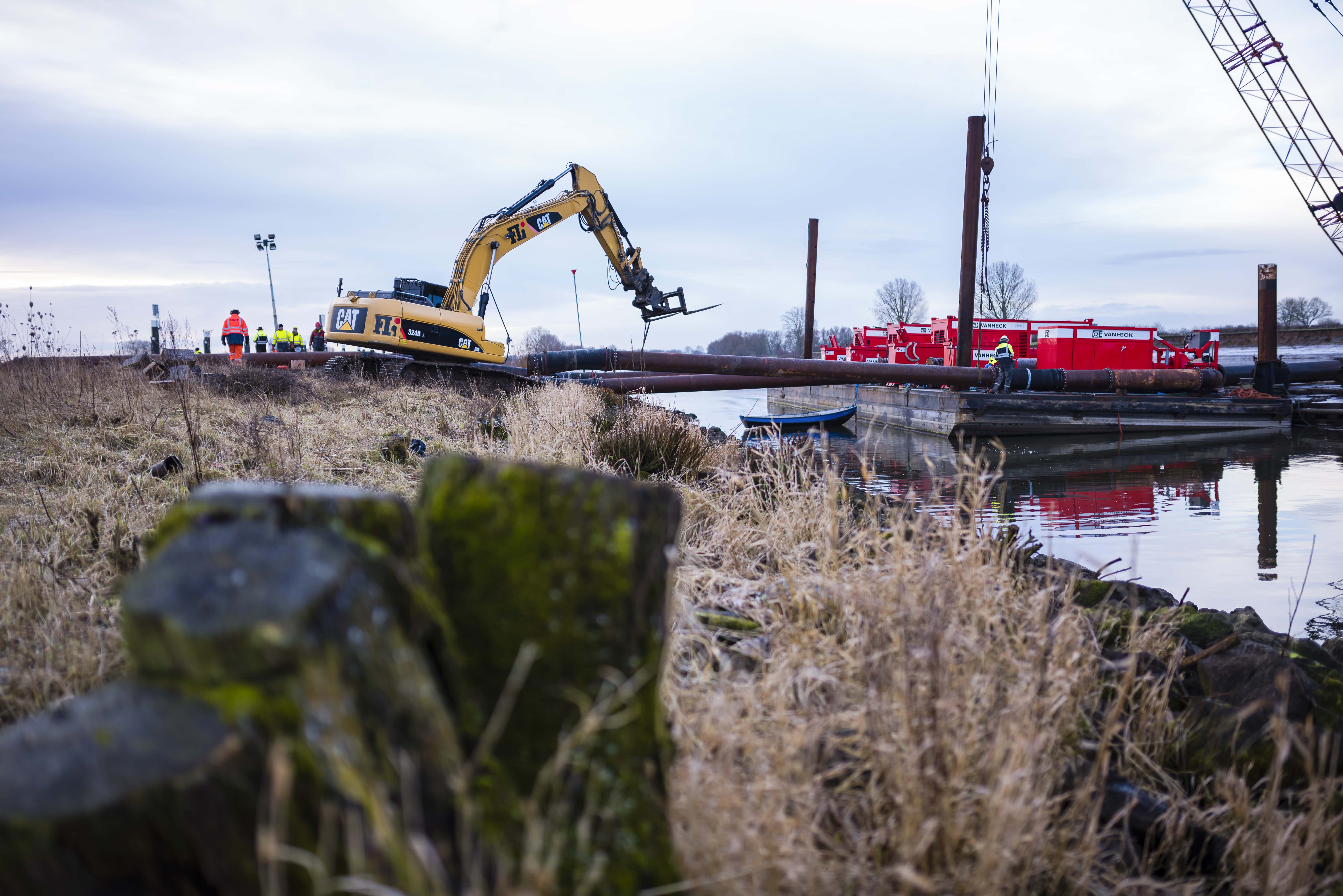 Opbouw van noodpompstation Werkzaamheden met een gele graafmachine en een ponton in het water met daarop rode pompcontainers en mannen in veiligheidskleding.