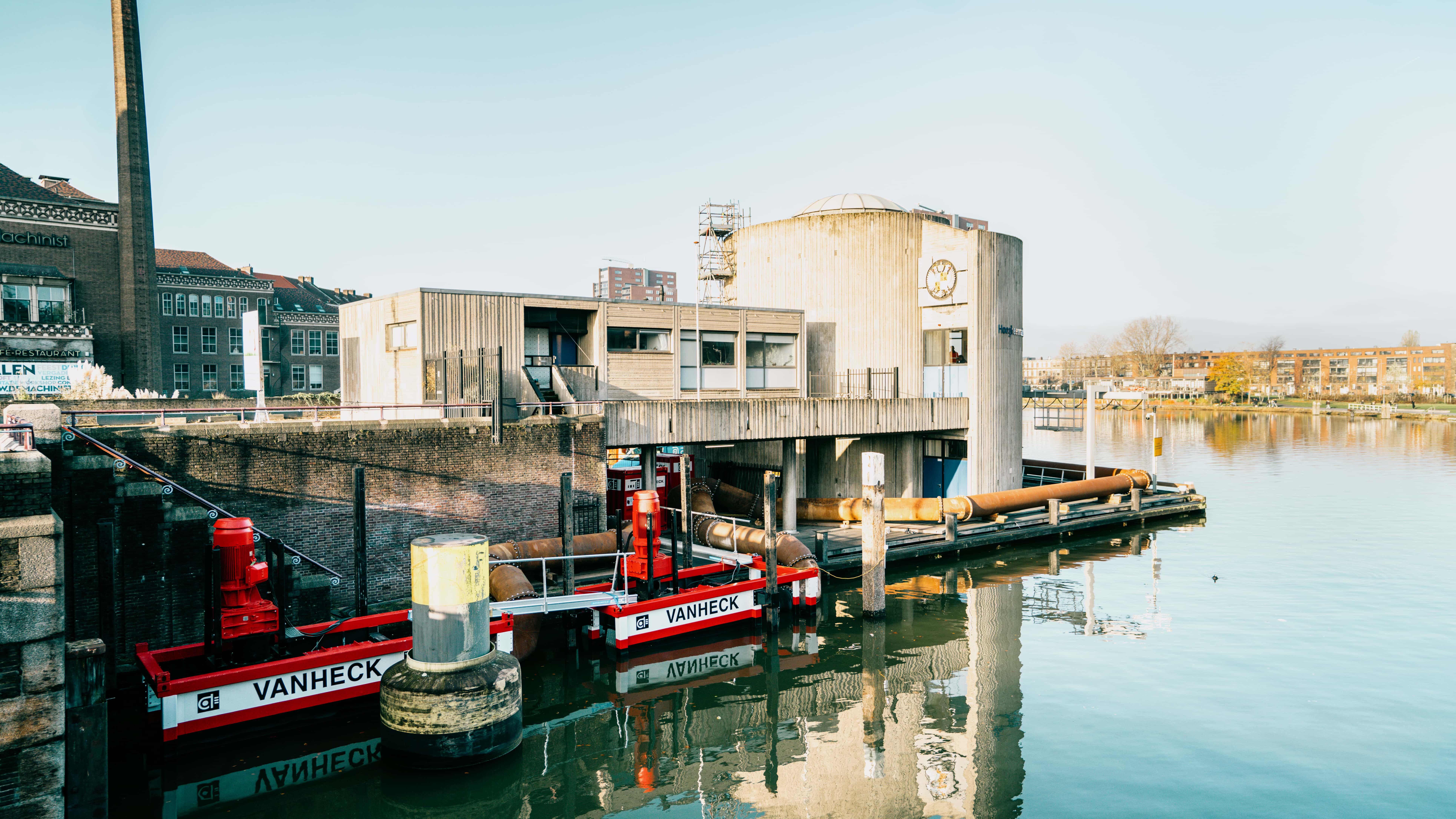 Gemaal Parksluizen Industrial waterfront scene with a concrete building, red waterpumps labeled "Vanheck," and calm reflective water under a clear sky.