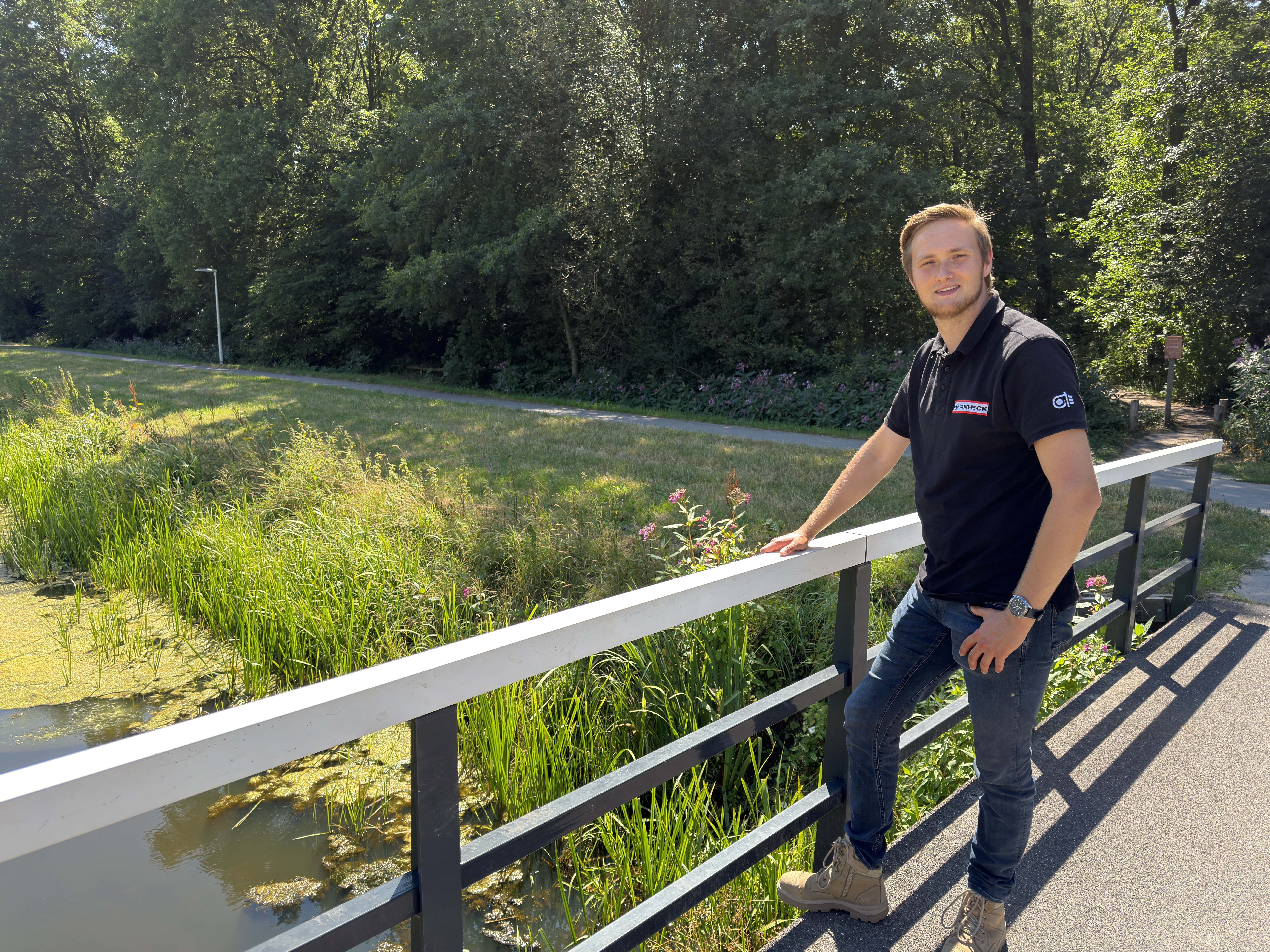 Een man staat op een brug naast een groen landschap met bomen en gras. Hij draagt een zwart T-shirt en lacht naar de camera.