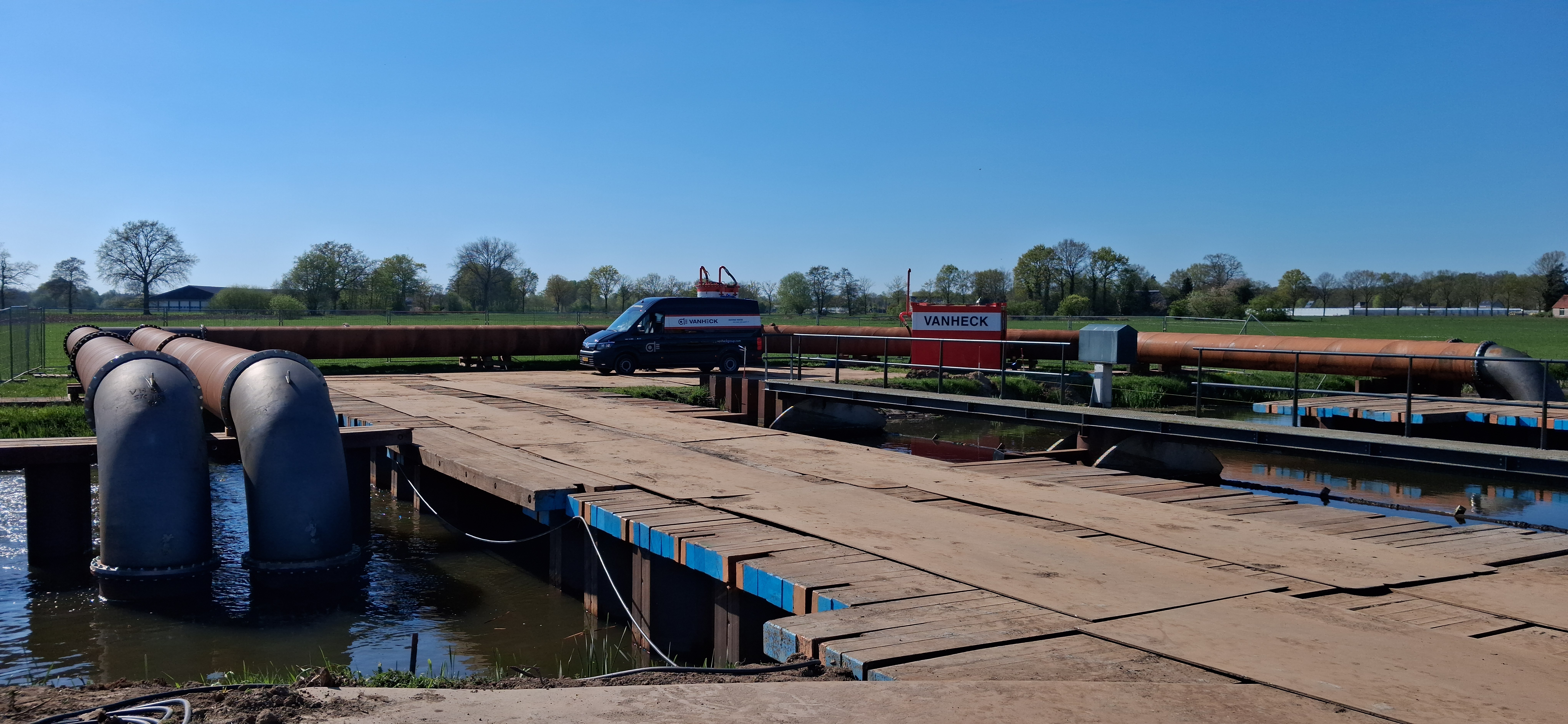 Hevelinstallatie bij een stuw Stalen leidingwerk dat tijdelijk water omleidt op een bouwplaats van rijplaten en draglineschotten onder een blauwe lucht. op de achtergrond staat een bedrijfsbus en een rode container met het opschrift "Van Heck"