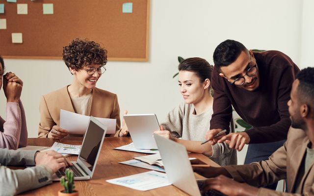 Large team collaborates at table with laptops and documents in modern office workspace.