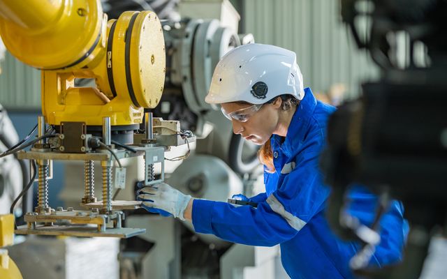 Female engineer in blue uniform and helmet operates industrial robot arm in a factory setting.