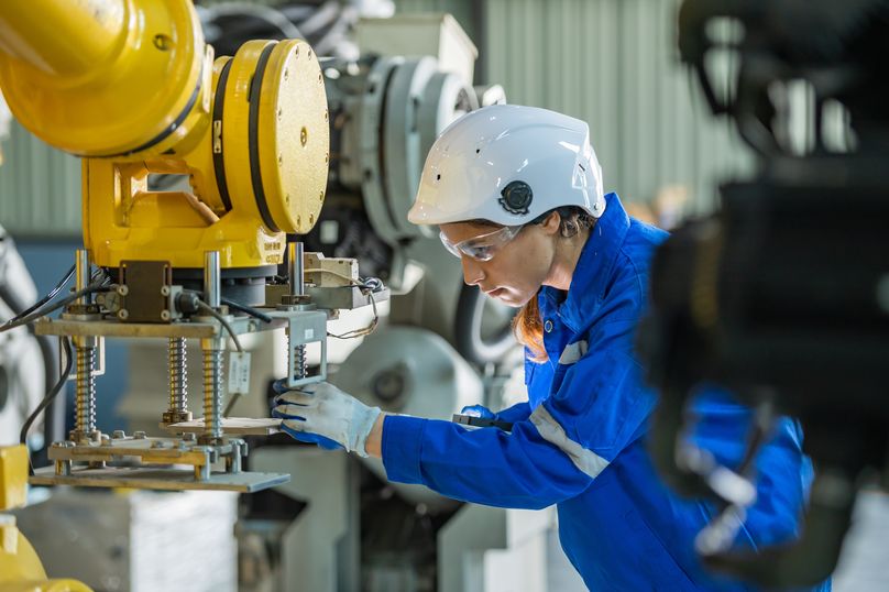 Female engineer in blue uniform and helmet operates industrial robot arm in a factory setting.