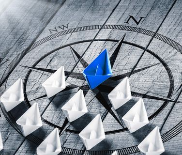 Paper boats on a compass, one blue leading white boats, symbolizing leadership and direction.